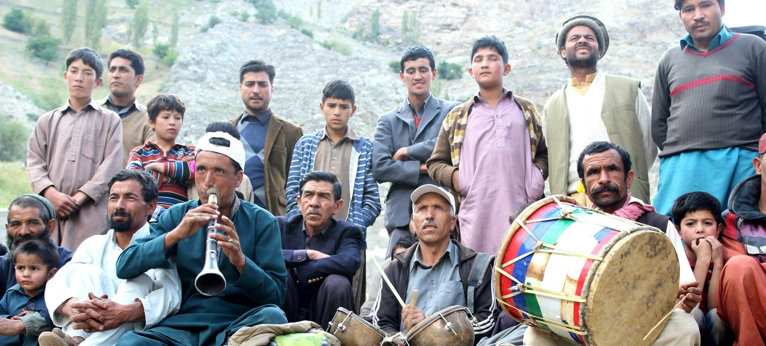 A group of men and children gathered outdoors, some playing musical instruments like a drum and a wind instrument, with mountains in the background.