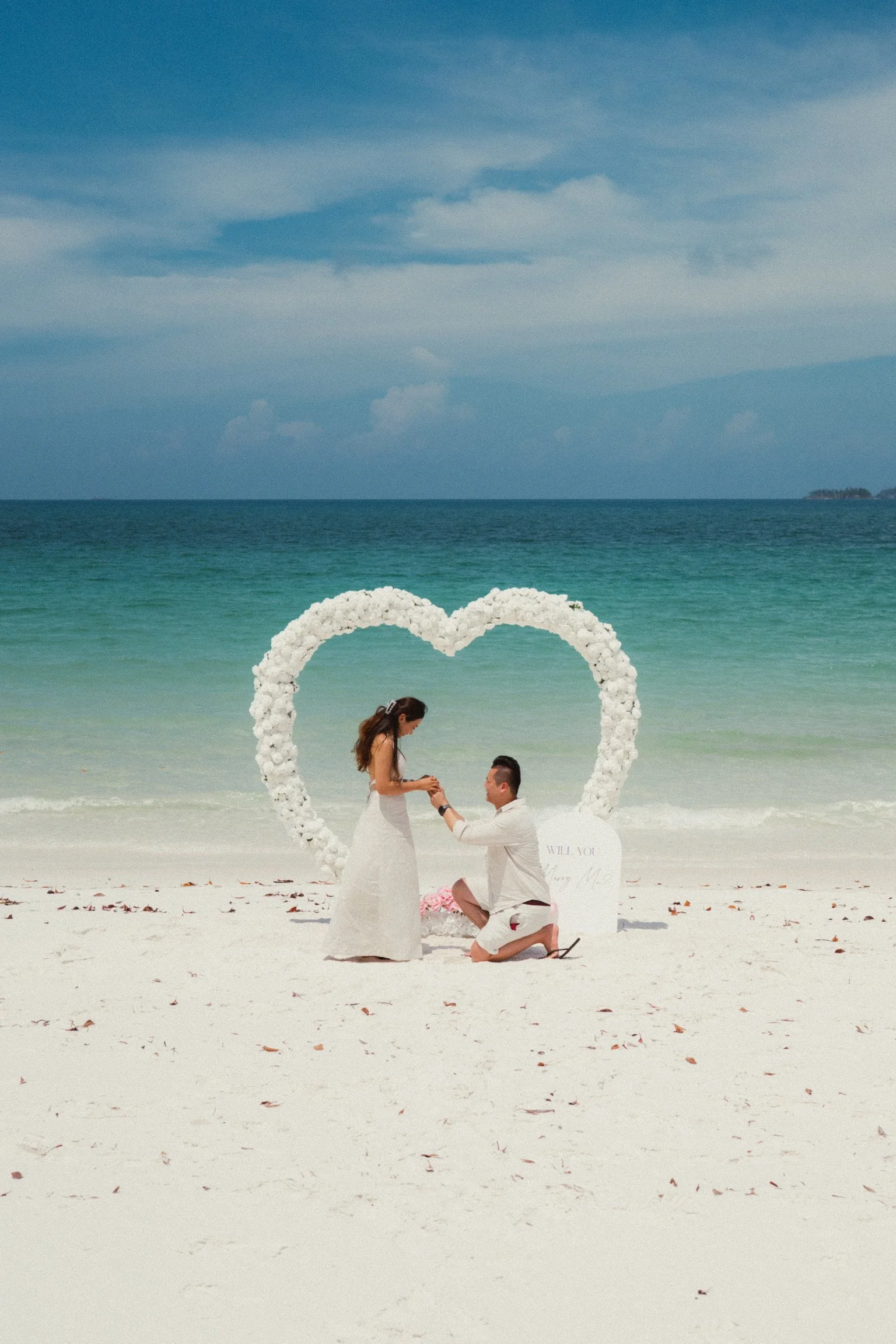 Luxury beach proposal setup in Airlie Beach and the Whitsundays featuring a white floral heart arch, romantic styling and oceanfront engagement experience by Salt & Silk Collective