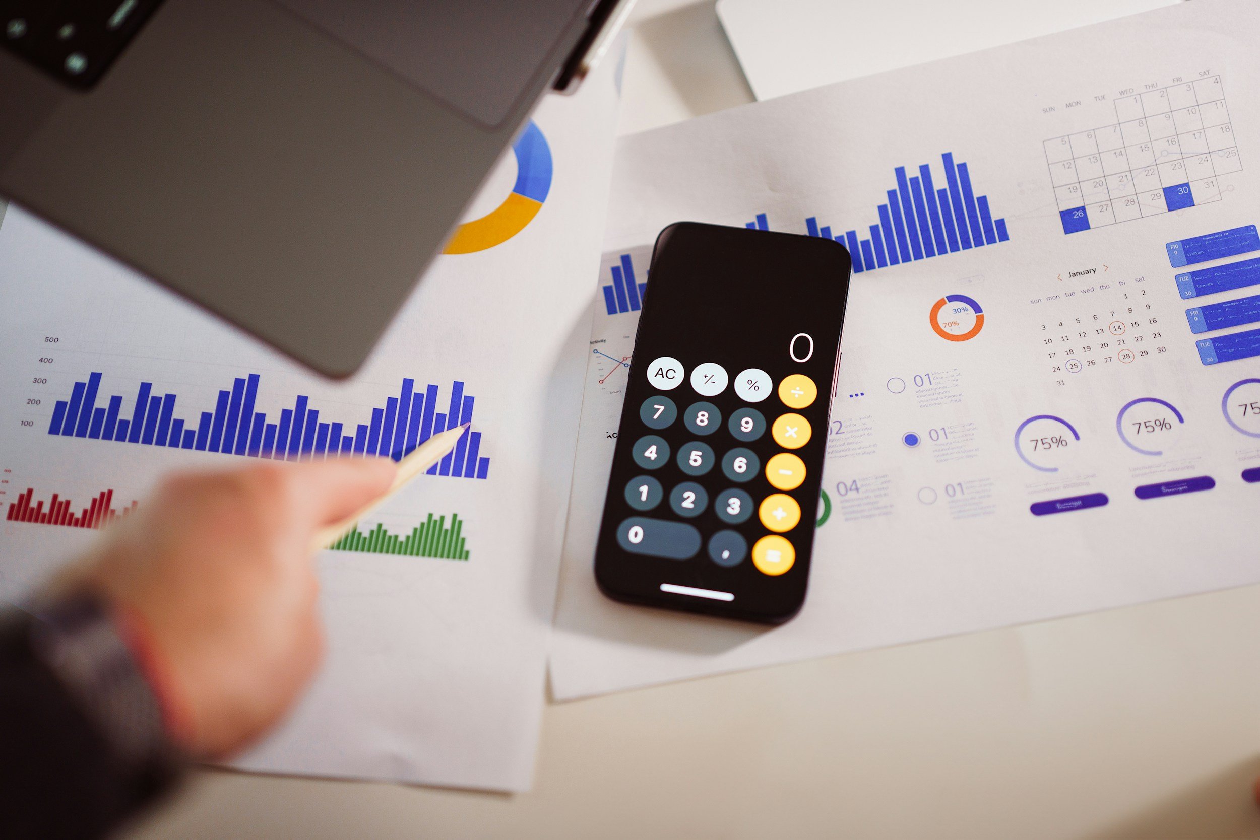 Desk with various financial charts including bar graphs and pie charts, a calculator, a smartphone displaying a calculator app, and a partially visible laptop.