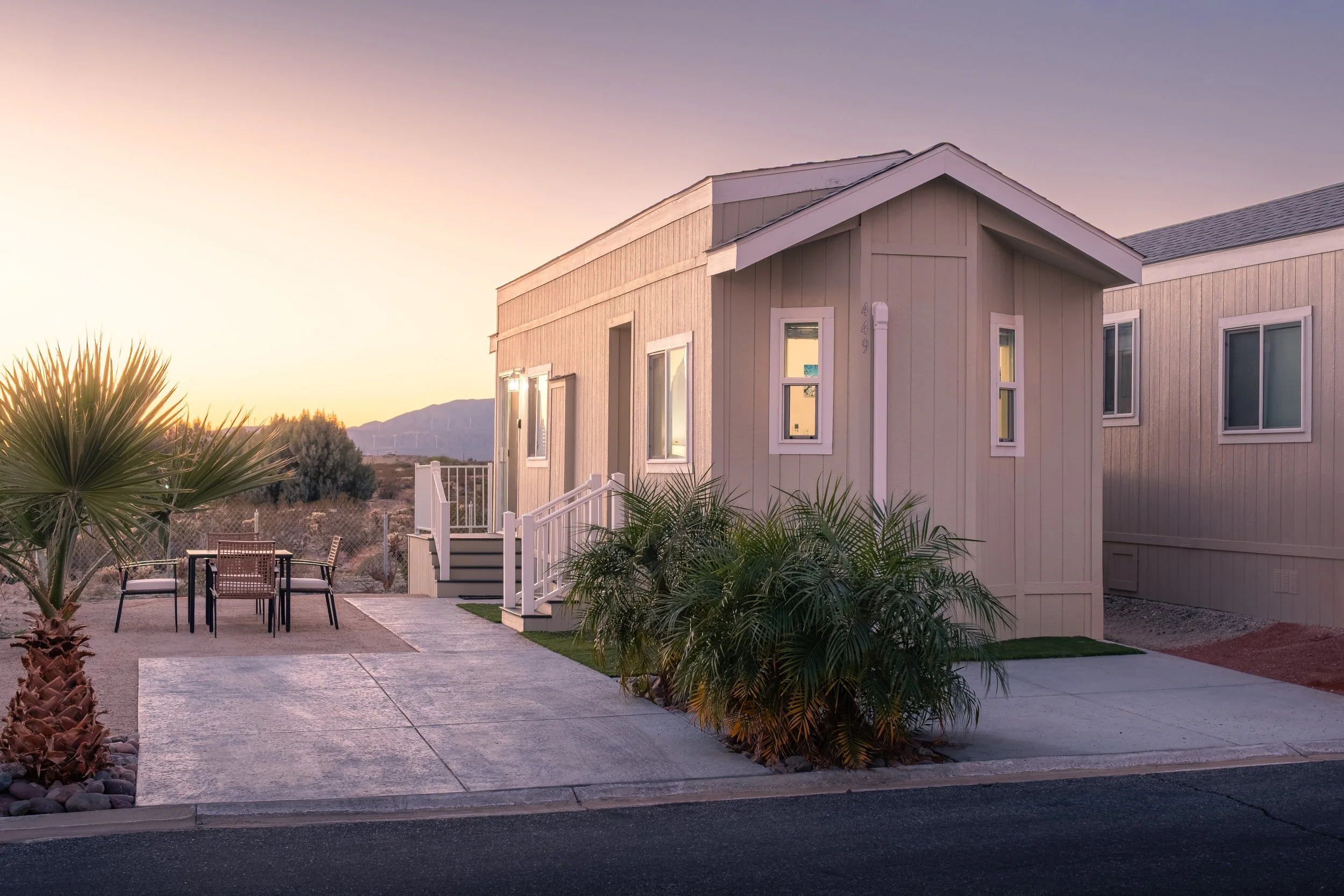 A residential house with beige vertical wood siding, white trim, multiple windows, and a covered entrance. There is a small outdoor seating area with a table and chairs on the sidewalk, surrounded by desert plants and rocks. The background shows a mountain range and a clear sky during sunset or sunrise.