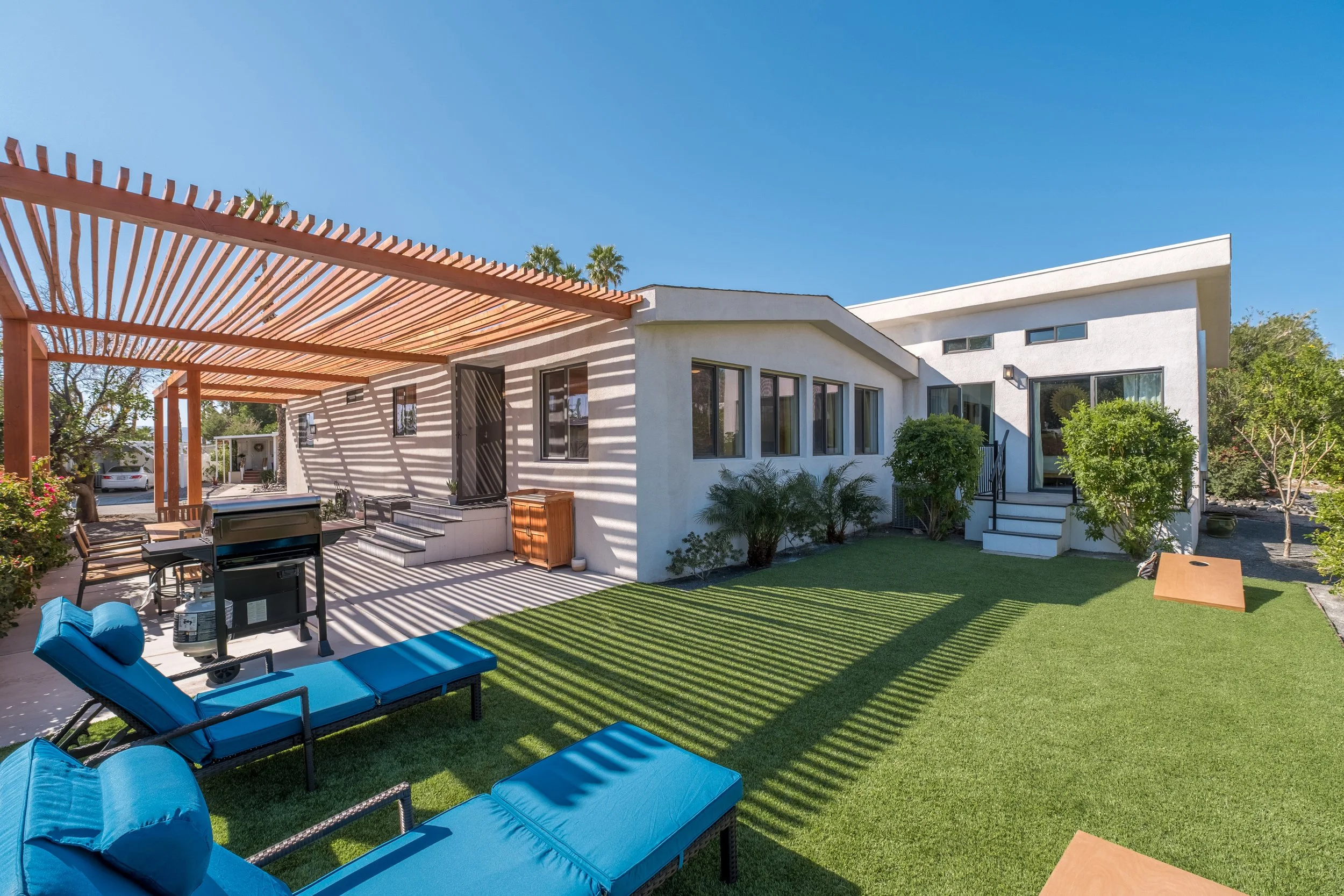 Modern white house with large windows and a backyard patio featuring blue lounge chairs, a grill, a pergola with wooden slats, and lush green bushes under a clear blue sky.