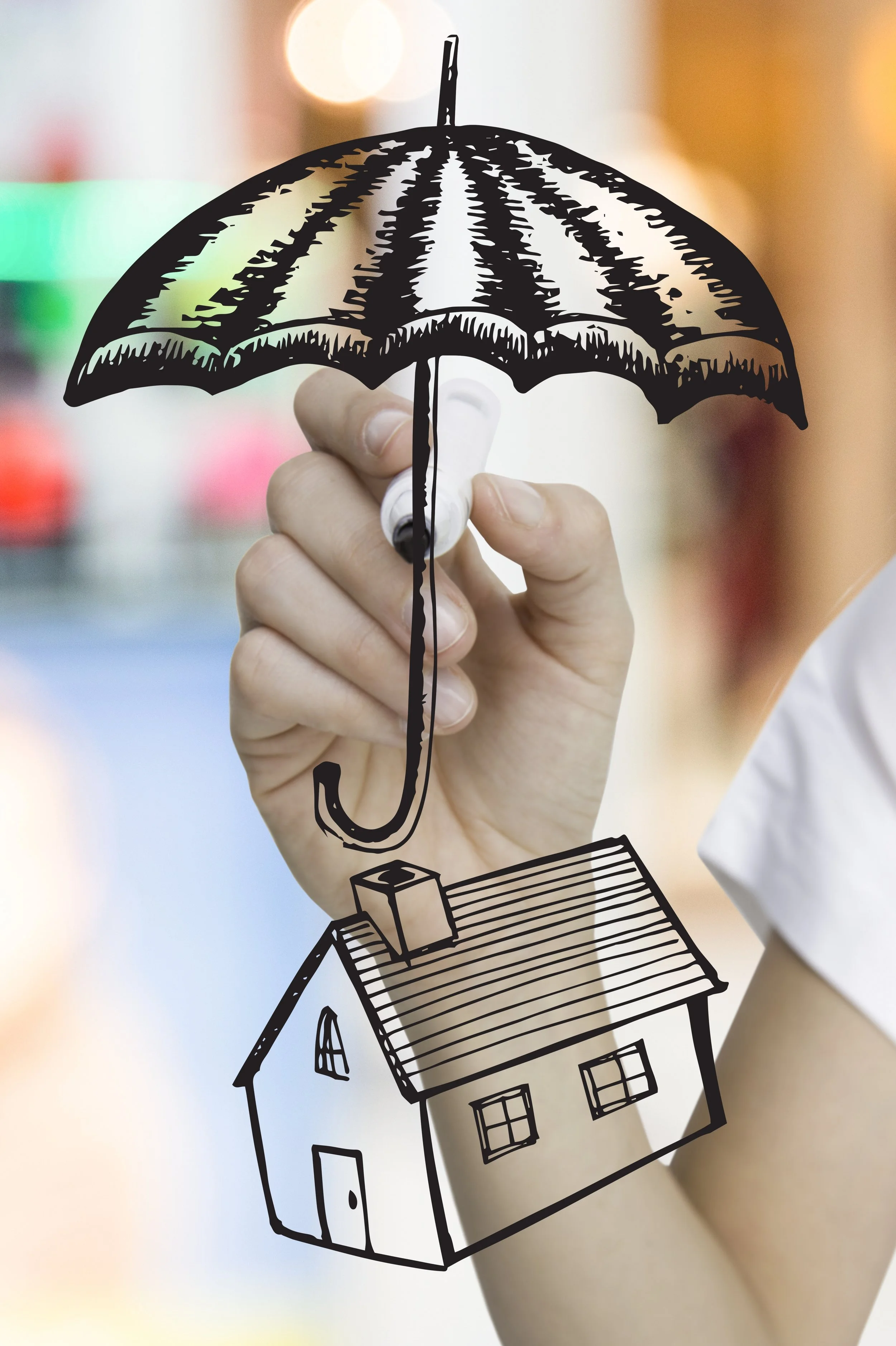 A hand holding a marker that draws a black outline of a house and an umbrella with a decorative roof, overlaid on a blurred background with warm tones.