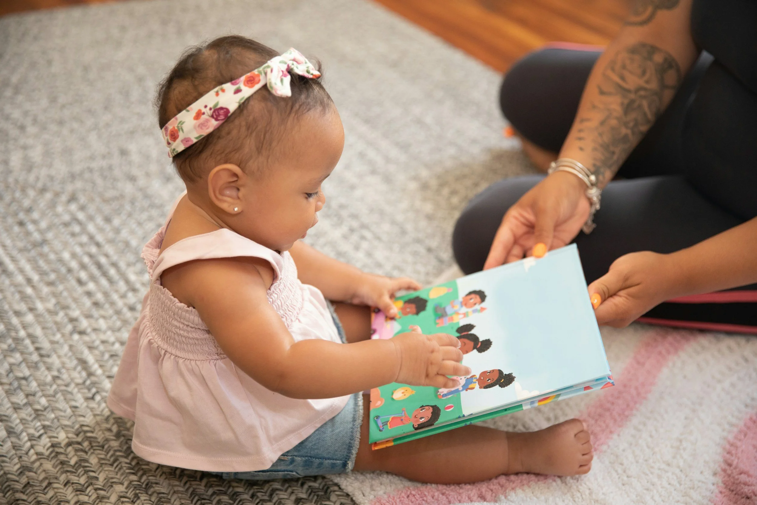 A young girl with a floral headband sitting on the floor, reading a colorful children's book with illustrations of children. An adult with tattoos is seated nearby, helping with the book.