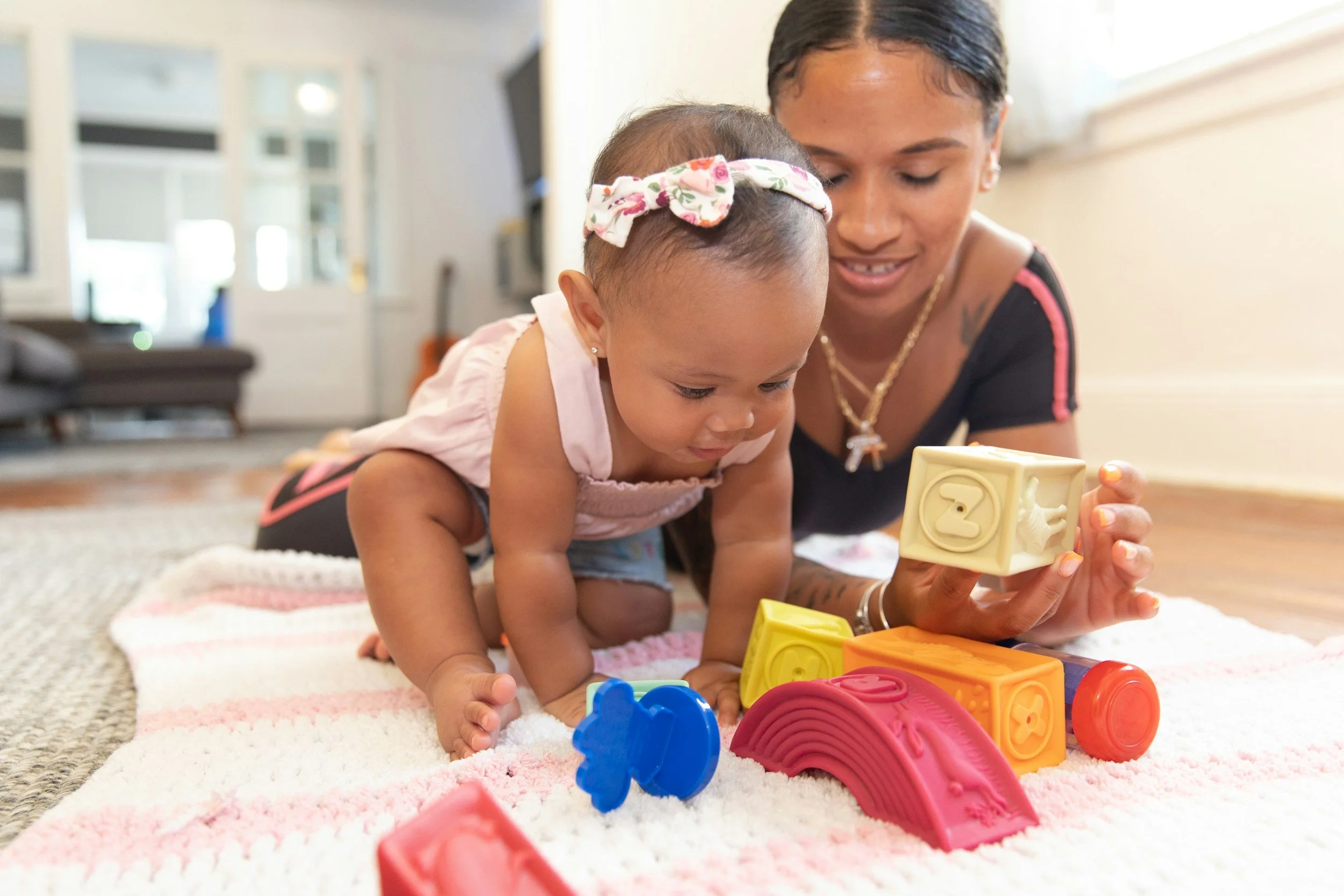 A woman and a young girl playing with colorful building blocks on a carpeted floor in a bright room.