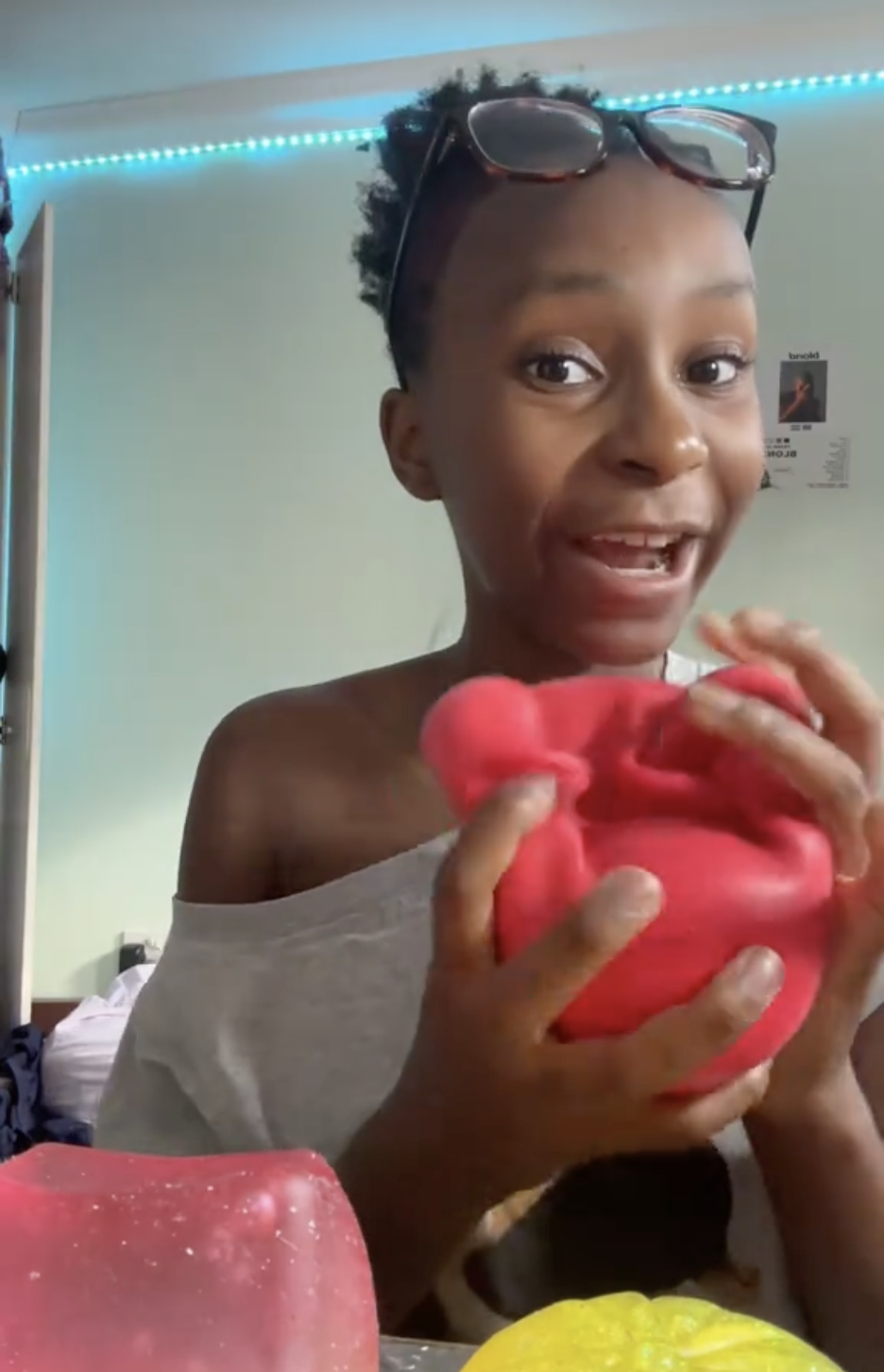 A young woman with glasses on her head smiling and holding a red slime in her hands, sitting in a room with light green walls and blue LED lighting.