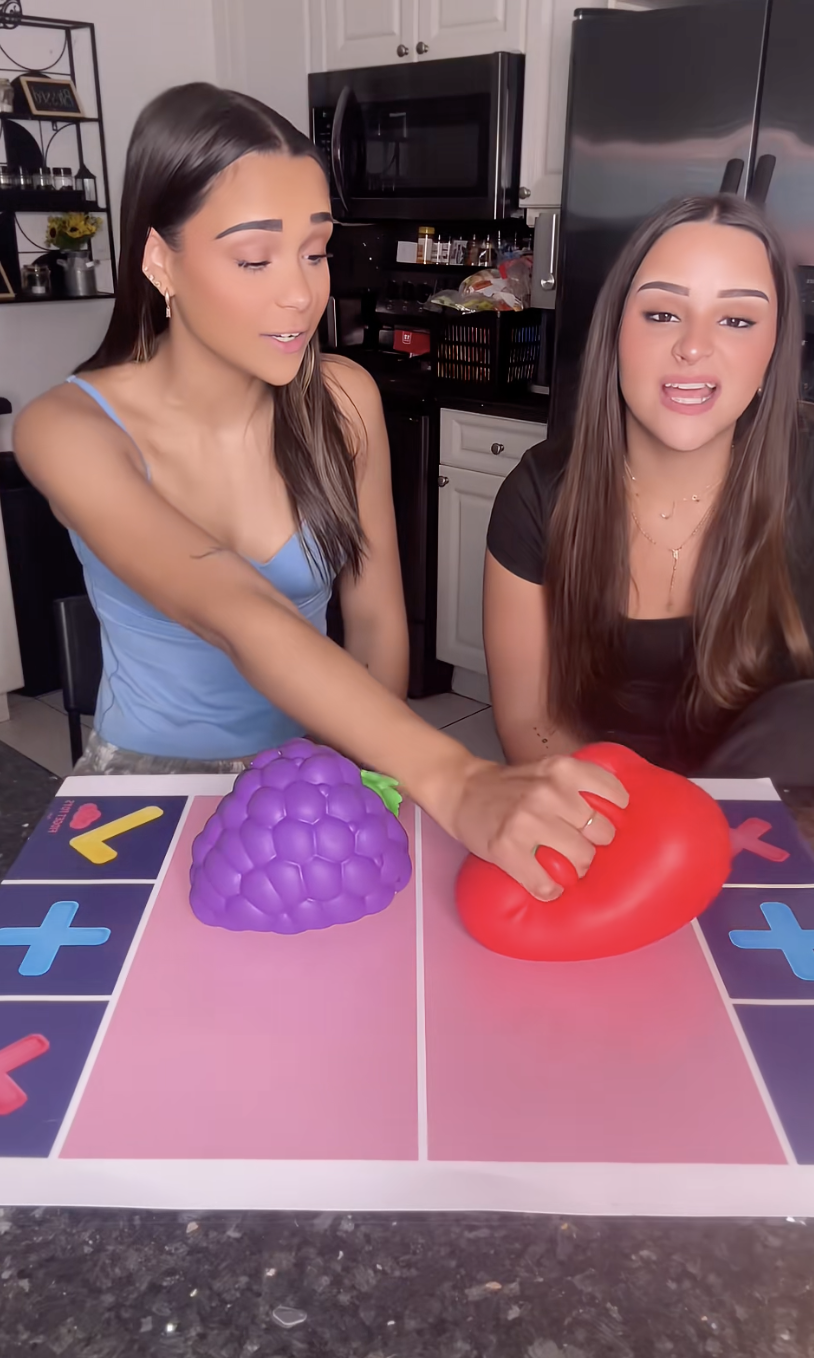 Two young women sitting at a table with game pieces, one purple shaped like a bunch of grapes and one red shaped like a tomato, in a kitchen setting.