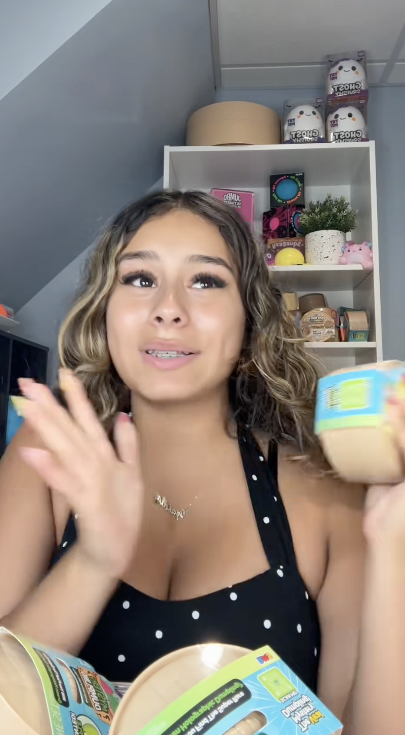 Young woman with curly hair holding a container of food, smiling, in front of a shelf with various decorative items and snacks.