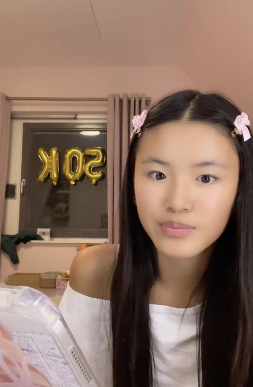 A young woman with long dark hair, wearing a white top with off-the-shoulder sleeves, pink hair clips, looking at the camera with a neutral expression. In the background, gold balloon letters spell out "SOP" inside a room with beige walls, a window with curtains, and some objects on a shelf.