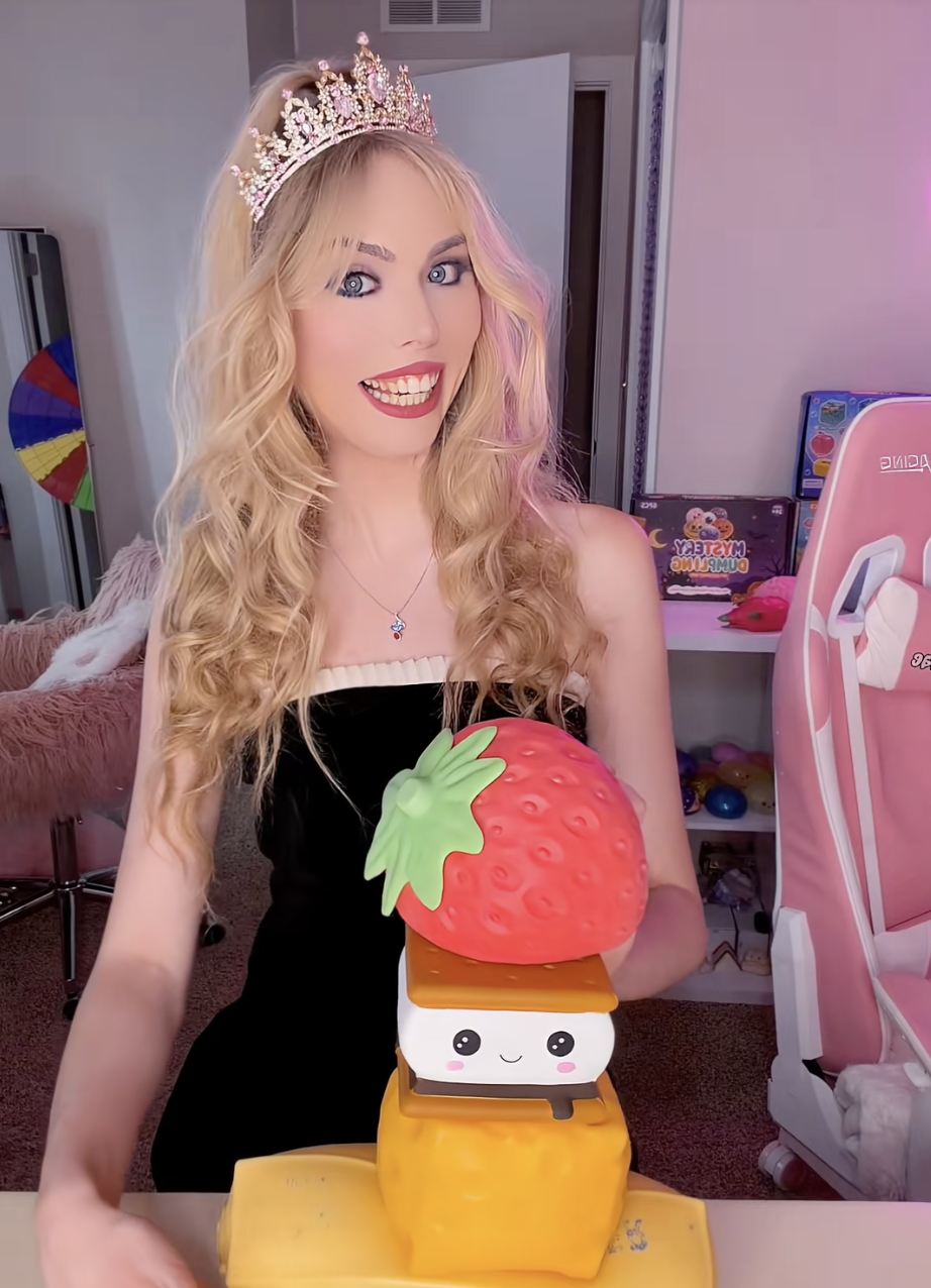 A young woman with long blonde curly hair wearing a tiara and black dress, smiling with a winking expression, holding a cute kawaii-style strawberry and pineapple-shaped cake with a smiling face, in a colorful room with toys and pink furniture.