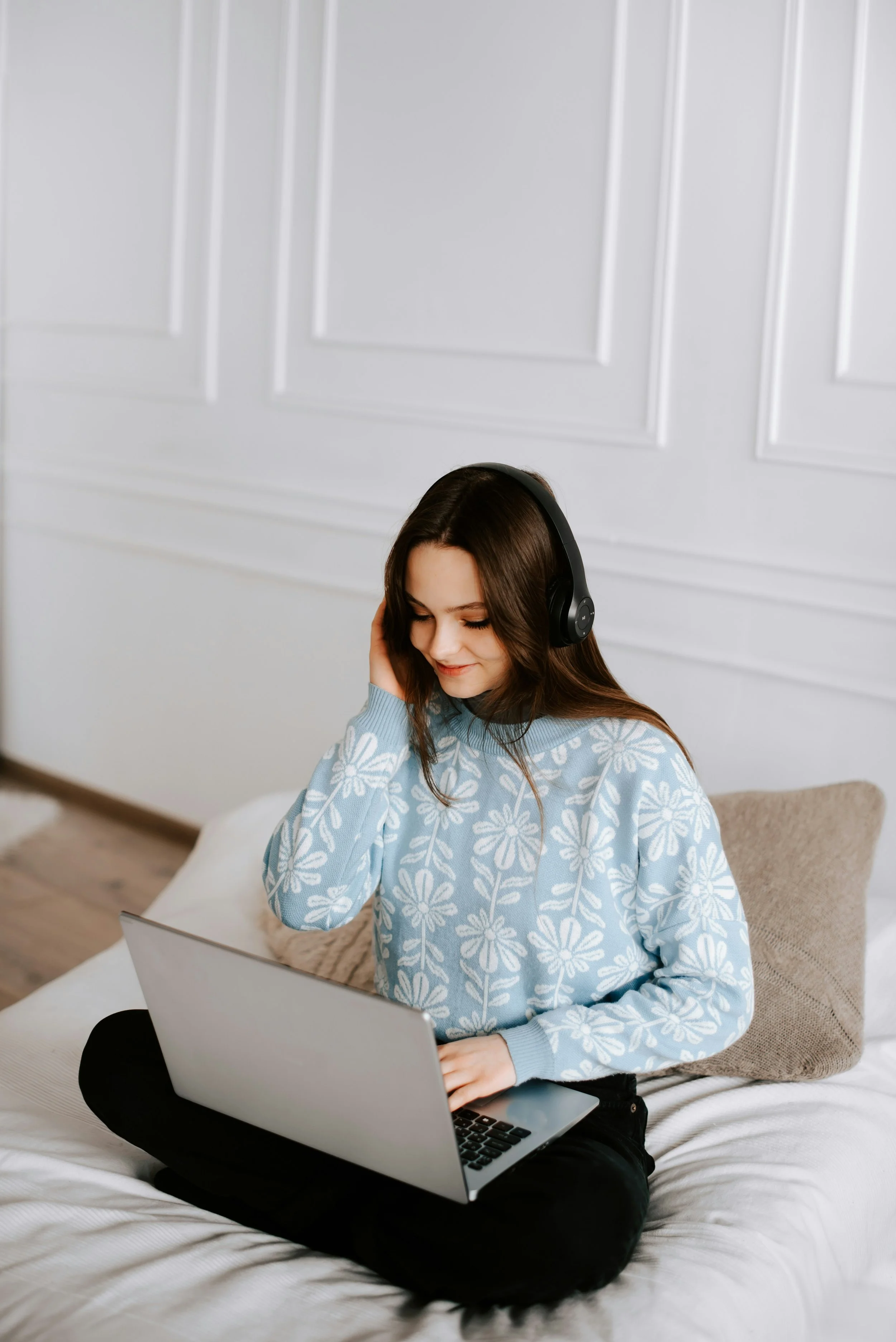 Young woman sitting on bed with laptop on lap, wearing headphones and a blue sweater with white floral pattern, smiling while working or studying at home.