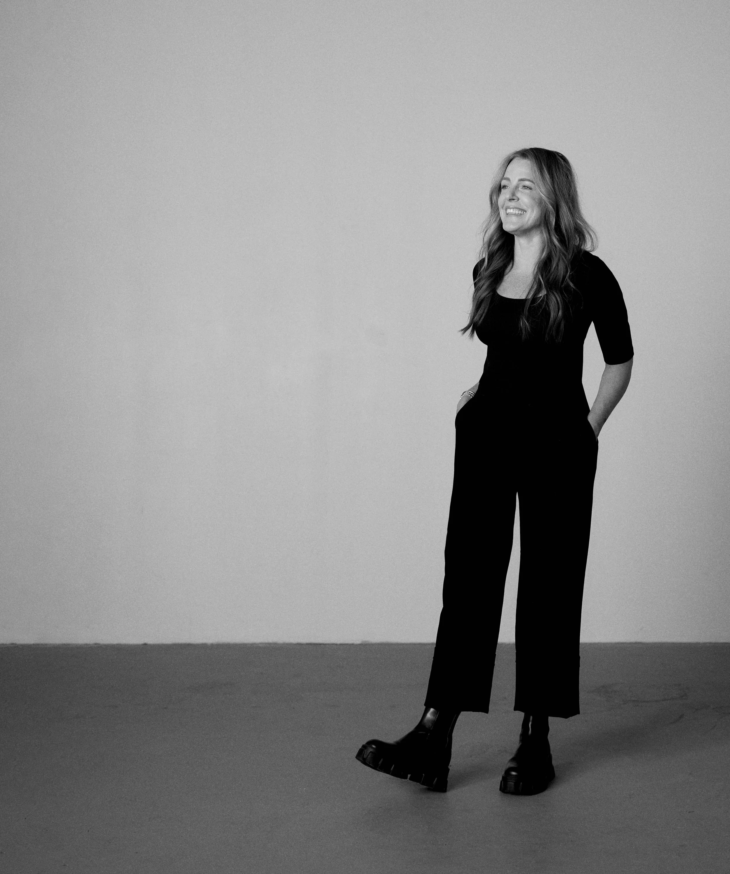 Black and white photo of a woman wearing high-heeled shoes, a long black dress, and a silver bracelet, standing on a beach with water in the background, smiling and looking to the side.