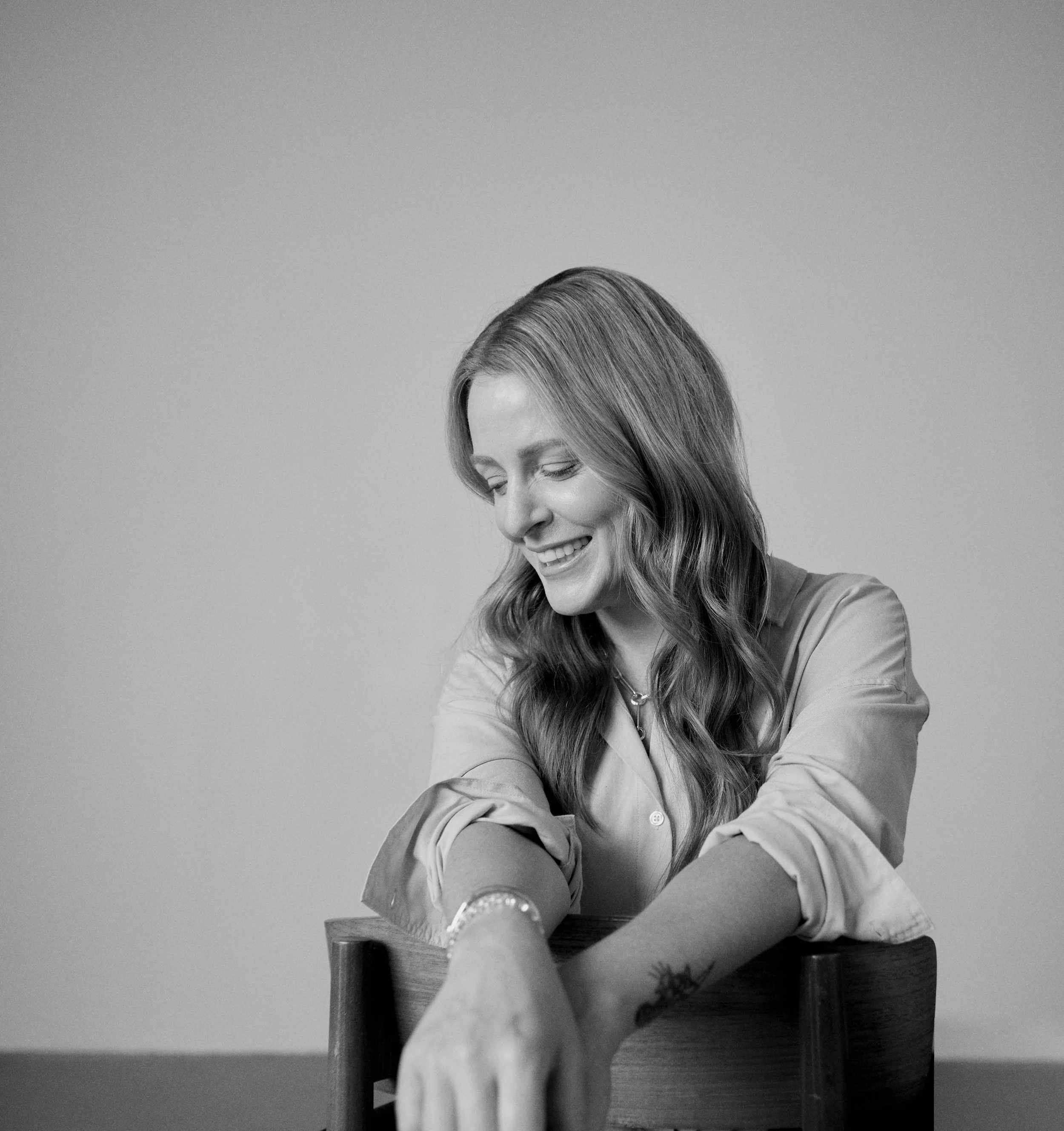 A woman with long hair smiling and looking down, sitting at a table with her left arm resting on a wooden chair, wearing a button-up shirt and a watch.