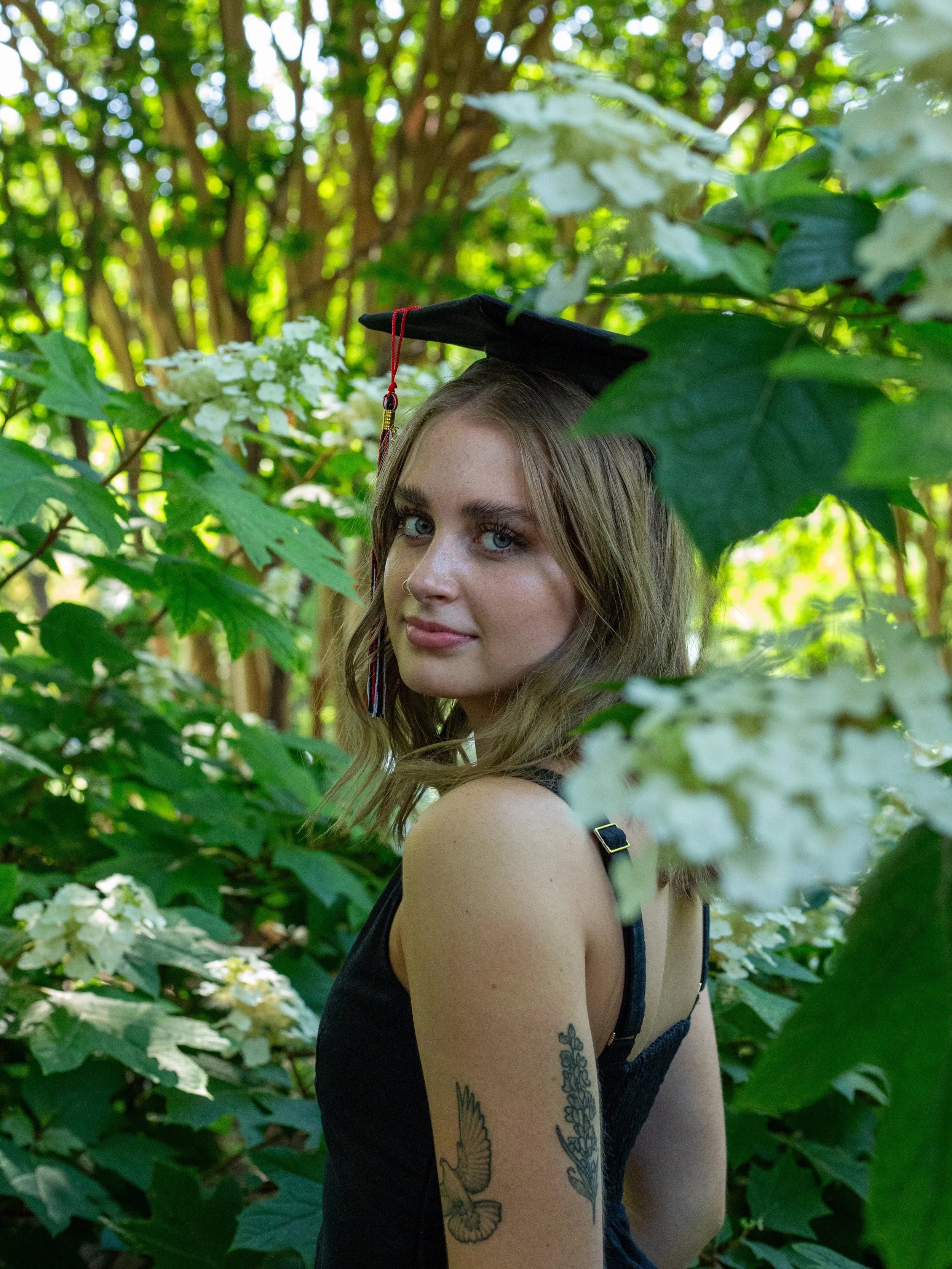 Young woman with long brown hair and tattoos on her arm, wearing a black sleeveless top, in a lush green outdoor setting with white flowers and large leaves.