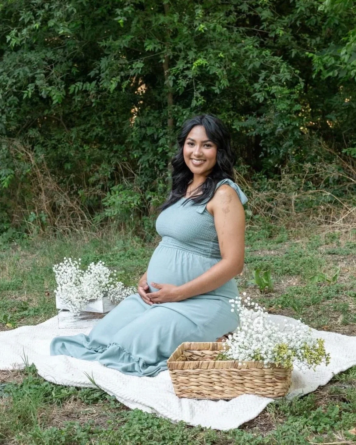 A pregnant woman sitting on a white blanket outdoors, surrounded by flowers and greenery, smiling at the camera.