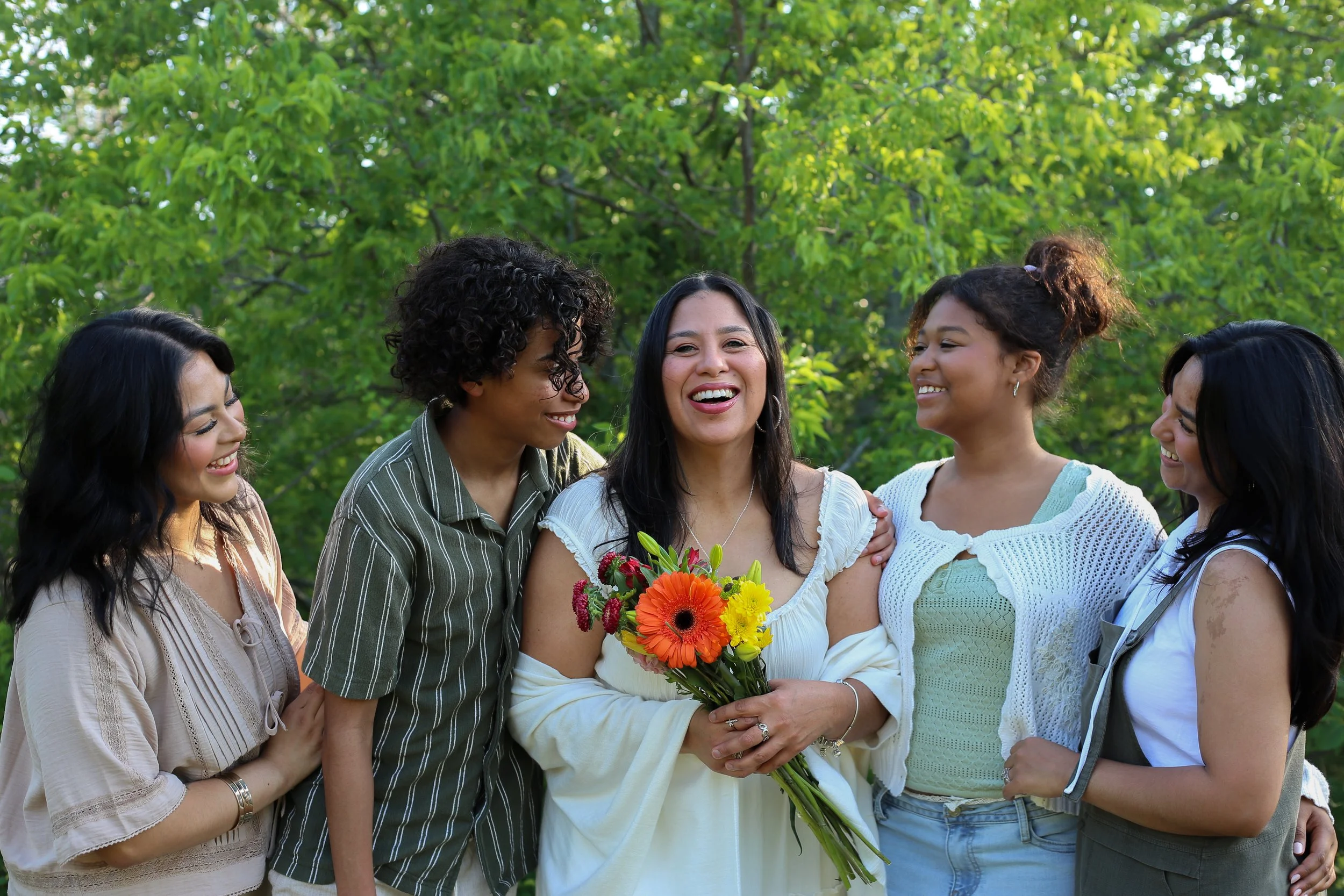 A group of women standing outdoors with lush green trees in the background, smiling and gathered around a woman holding a colorful bouquet of flowers.