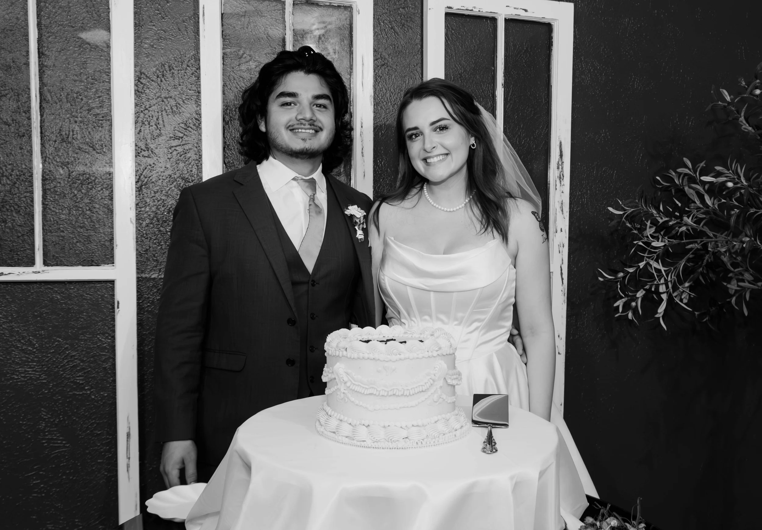A black and white photo of a young couple at their wedding, standing behind a table with a wedding cake. The groom has shoulder-length dark hair and is wearing a suit and tie. The bride has long, wavy hair and is wearing a wedding dress, pearl necklace, and veil. They are smiling and standing close together, with a window and decorative plants in the background.