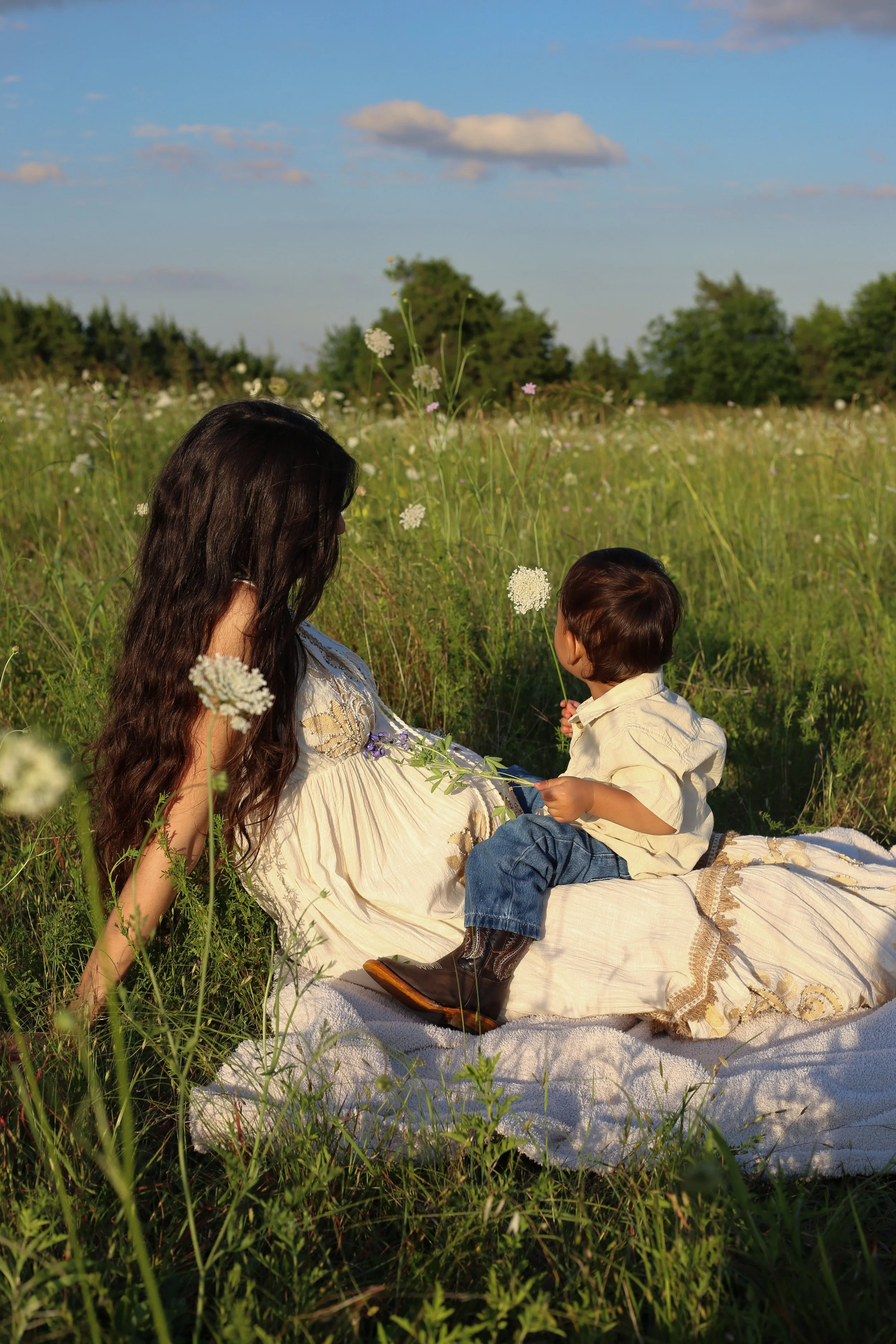 A woman and a young boy sitting on a white blanket in a grassy field with wildflowers, trees, and a partly cloudy sky in the background.