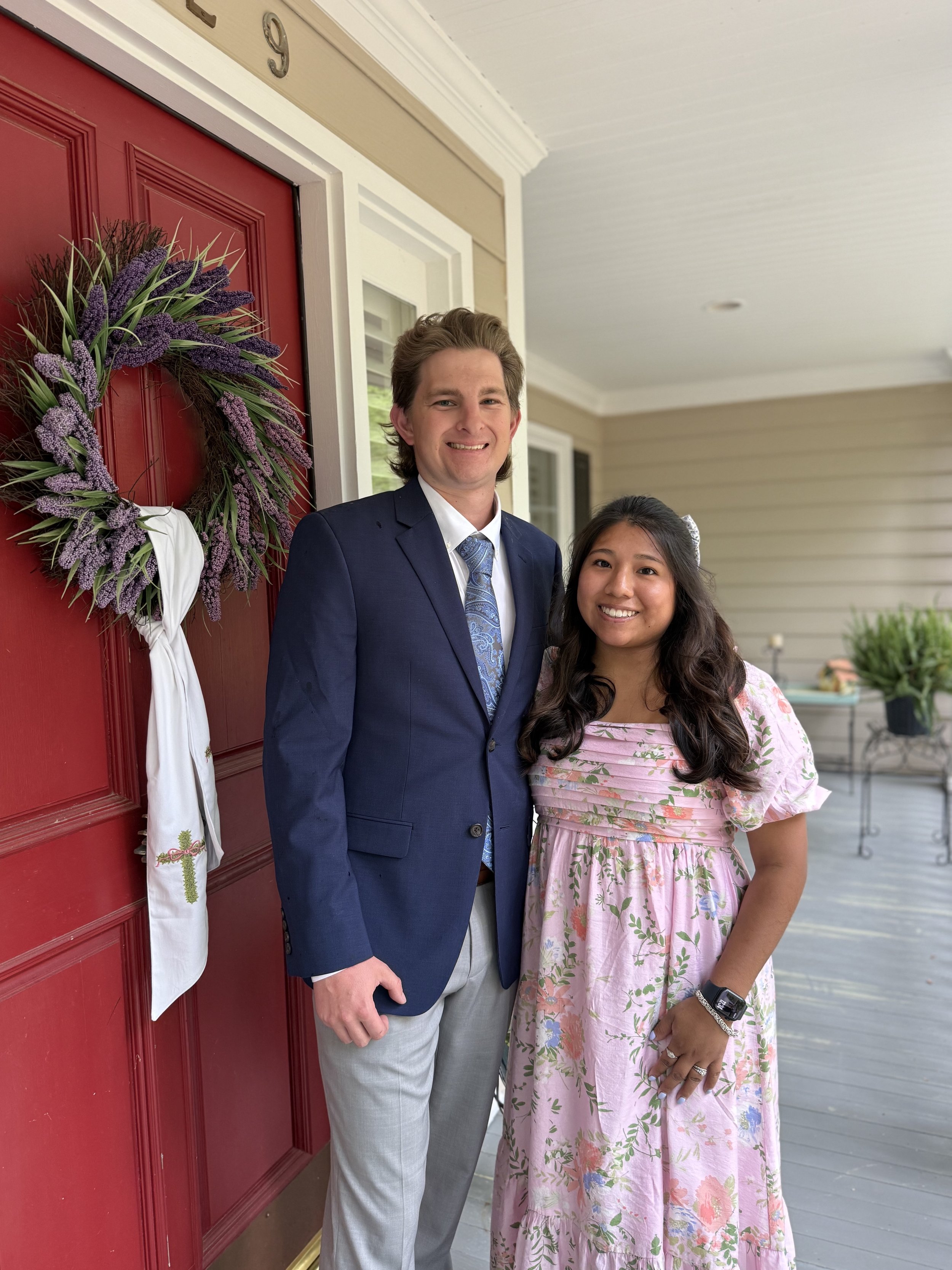 A young man in a navy blue suit with a patterned tie and a young woman in a pink floral dress standing in front of a red door decorated with a purple wreath and a white cloth tied with a green ornament. The door has the number 9 above it. They are smiling and standing close together.
