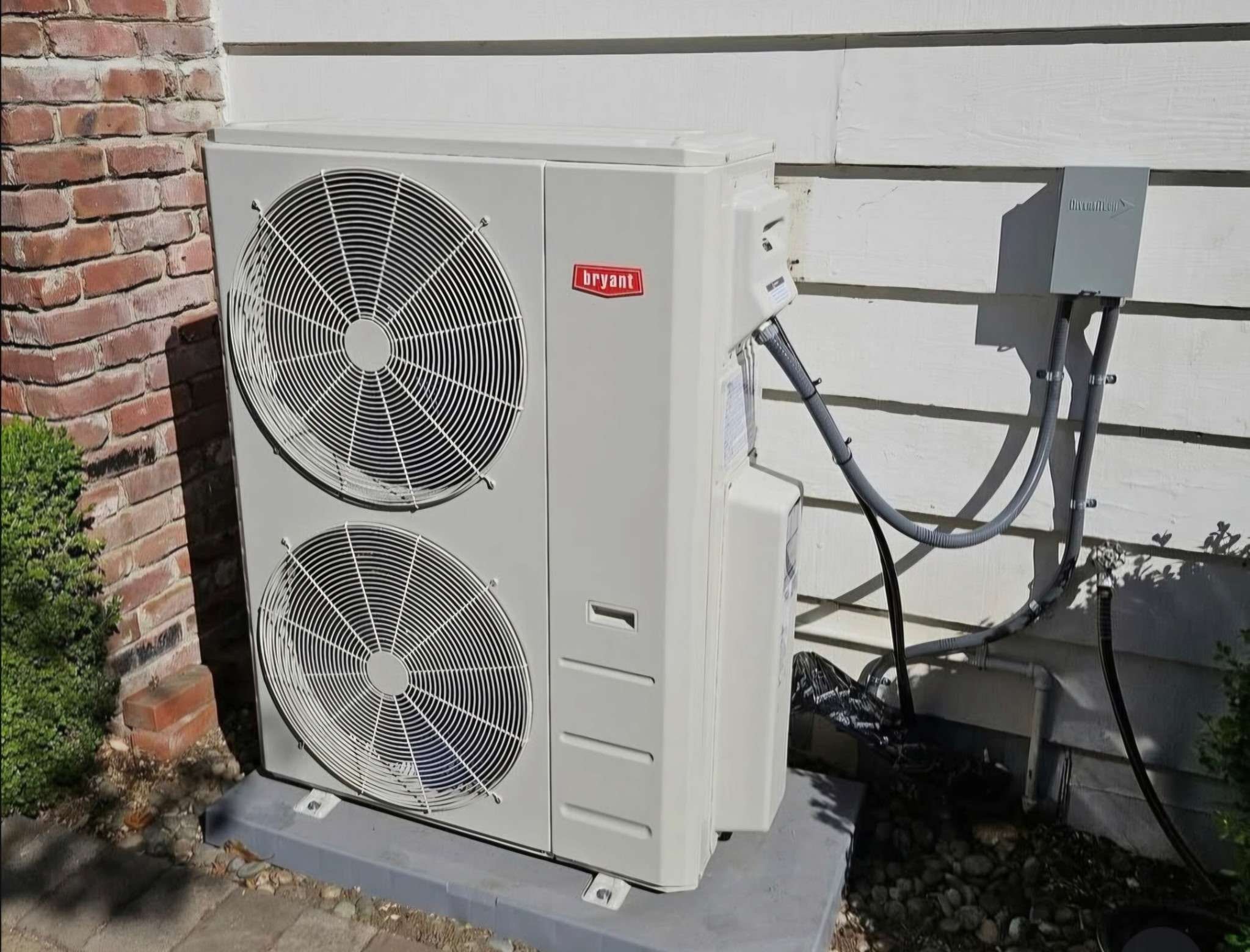 Exterior of an HVAC unit with Bryant logo, mounted on a stand outside a building with a brick wall on the left and a white wooden wall on the right, some electrical boxes and pipes connected to the unit.