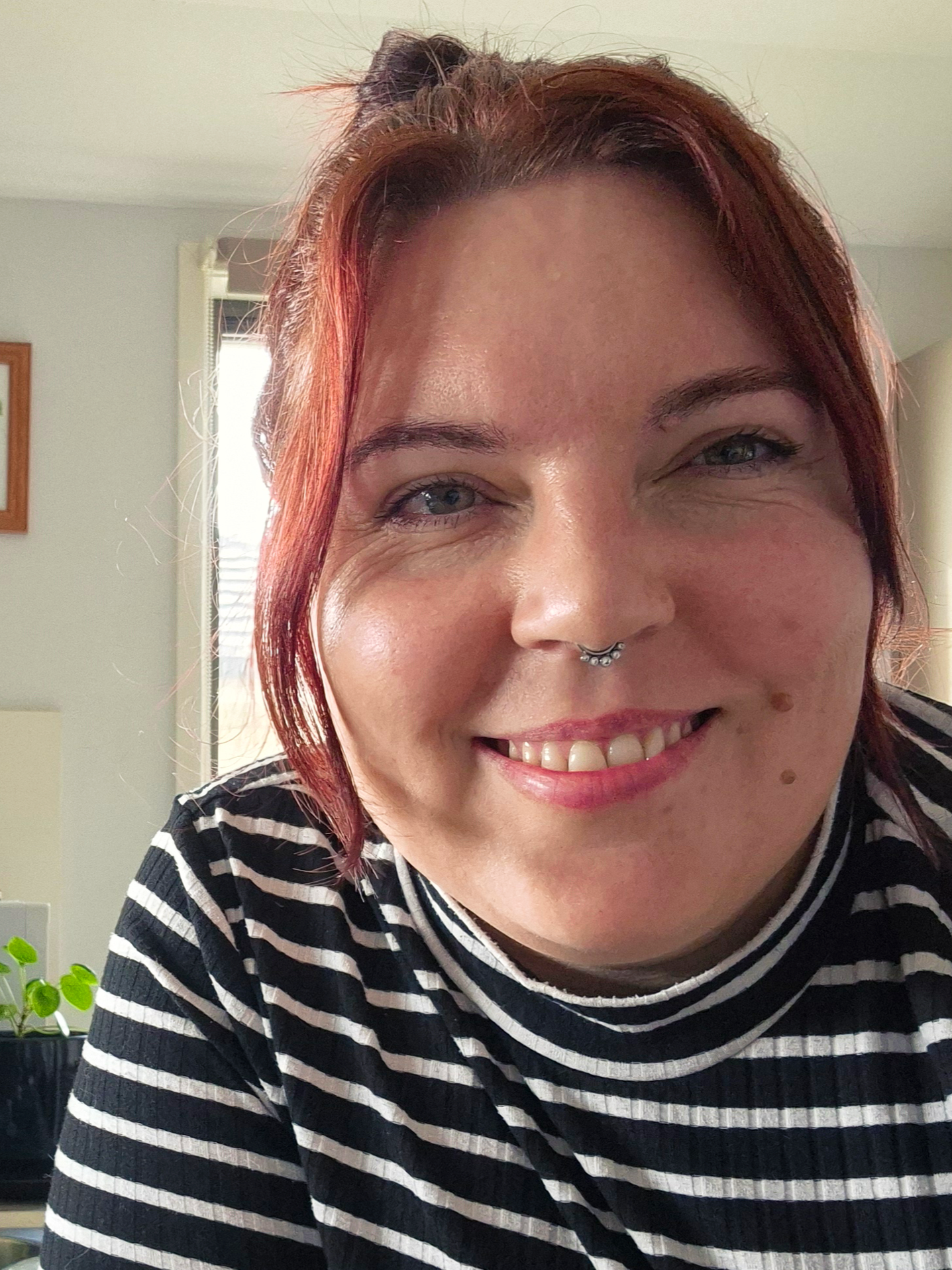 A smiling woman with red hair, hazel eyes, a septum piercing, wearing a black and white striped shirt, in a room with light-colored walls, a window, and a potted plant.