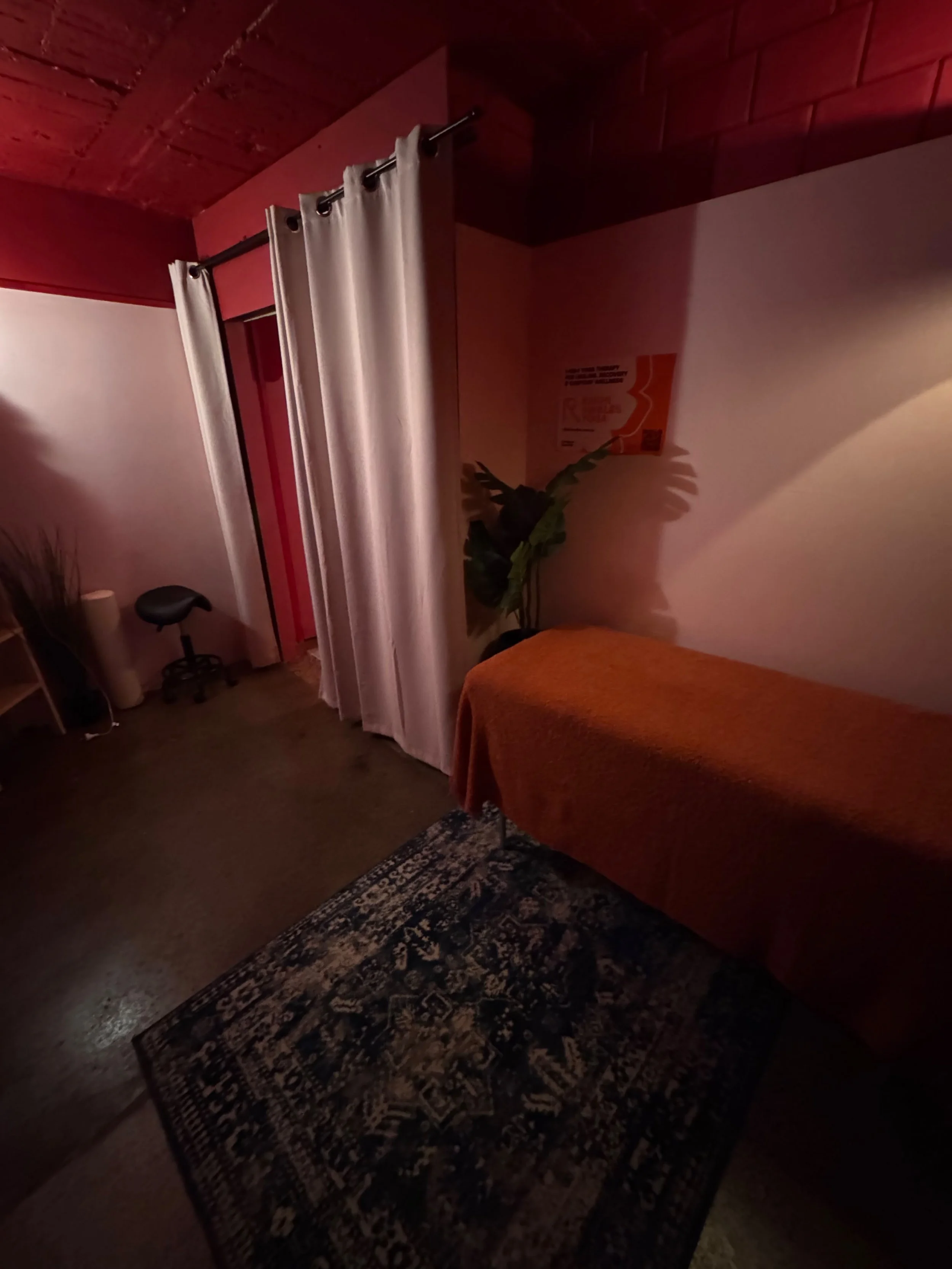 A dimly lit room with a dark patterned rug on a concrete floor, a bed with an orange cover, a green plant next to the bed, white curtains hanging from a black rod, and a small chair with wheels in the corner.