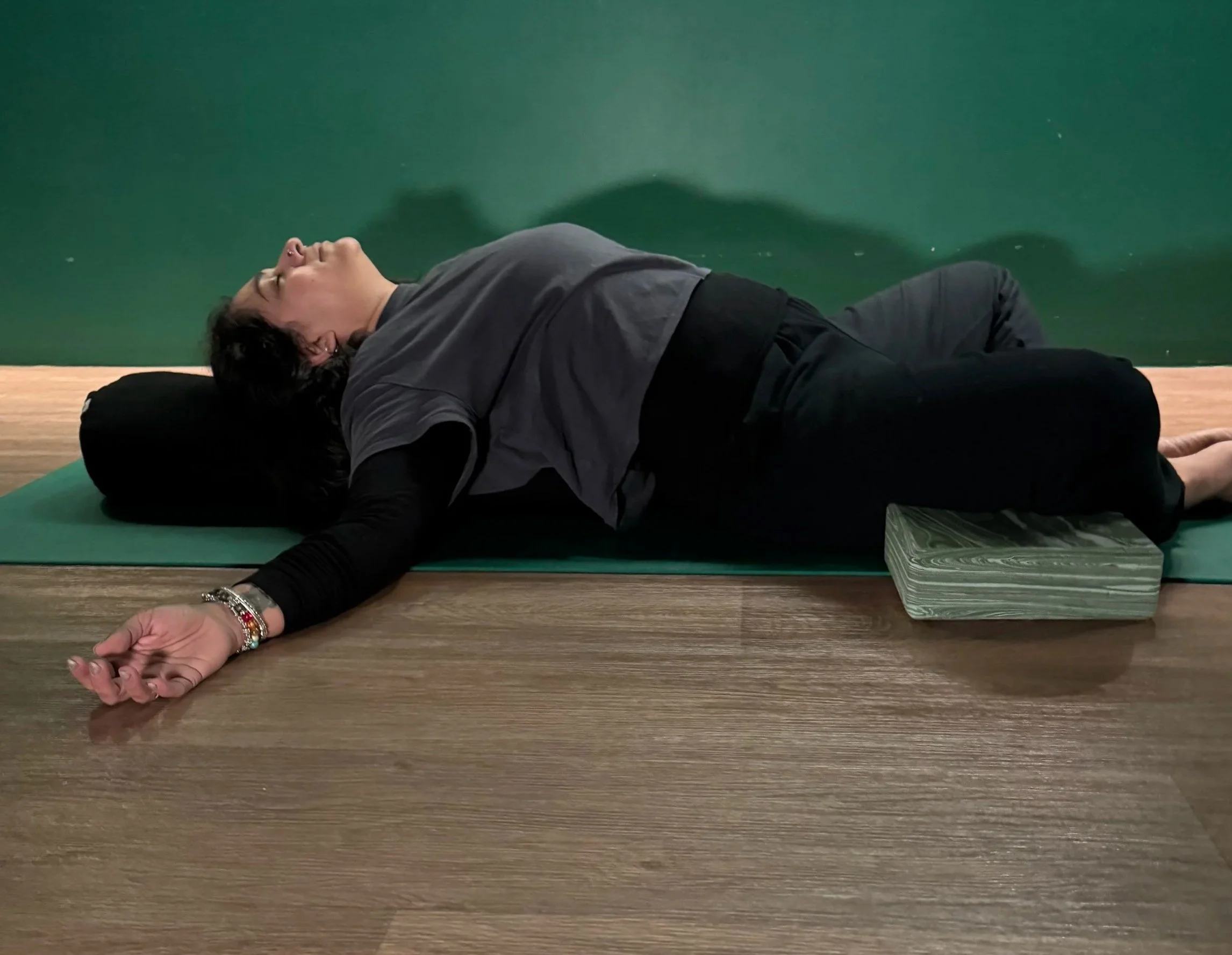 Person lying on a yoga mat with eyes closed, arm stretched out, and a stack of books nearby, against a dark green wall.