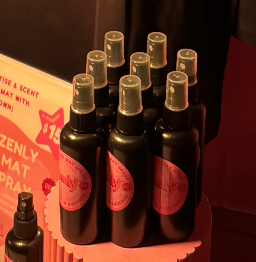 A display of small spray bottles with a pink label, arranged on a pink tray, with a partially visible sign on the left.