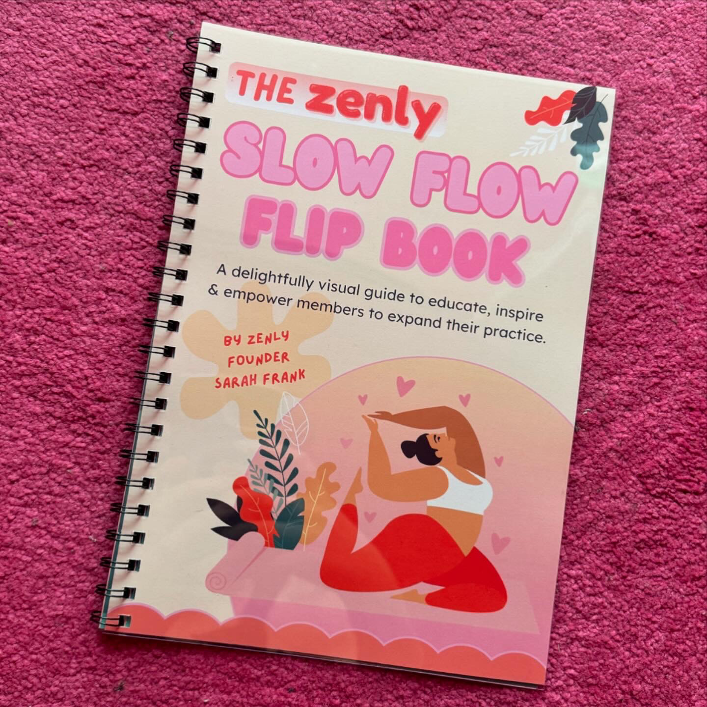 A spiral-bound book titled 'The Zenly Slow Flow Flip Book' placed on a pink textured surface. The book features an illustration of a person doing yoga surrounded by colorful leaves and hearts, with a description indicating it is a visual guide to educate, inspire, and empower members.