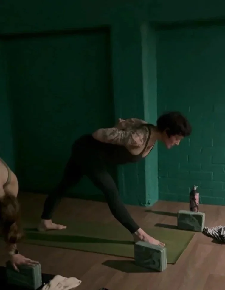 Woman in black leggings and a tank top doing yoga in a studio, reaching towards her left ankle with a yoga block in front of her, while an instructor or fellow participant extends a hand towards her.