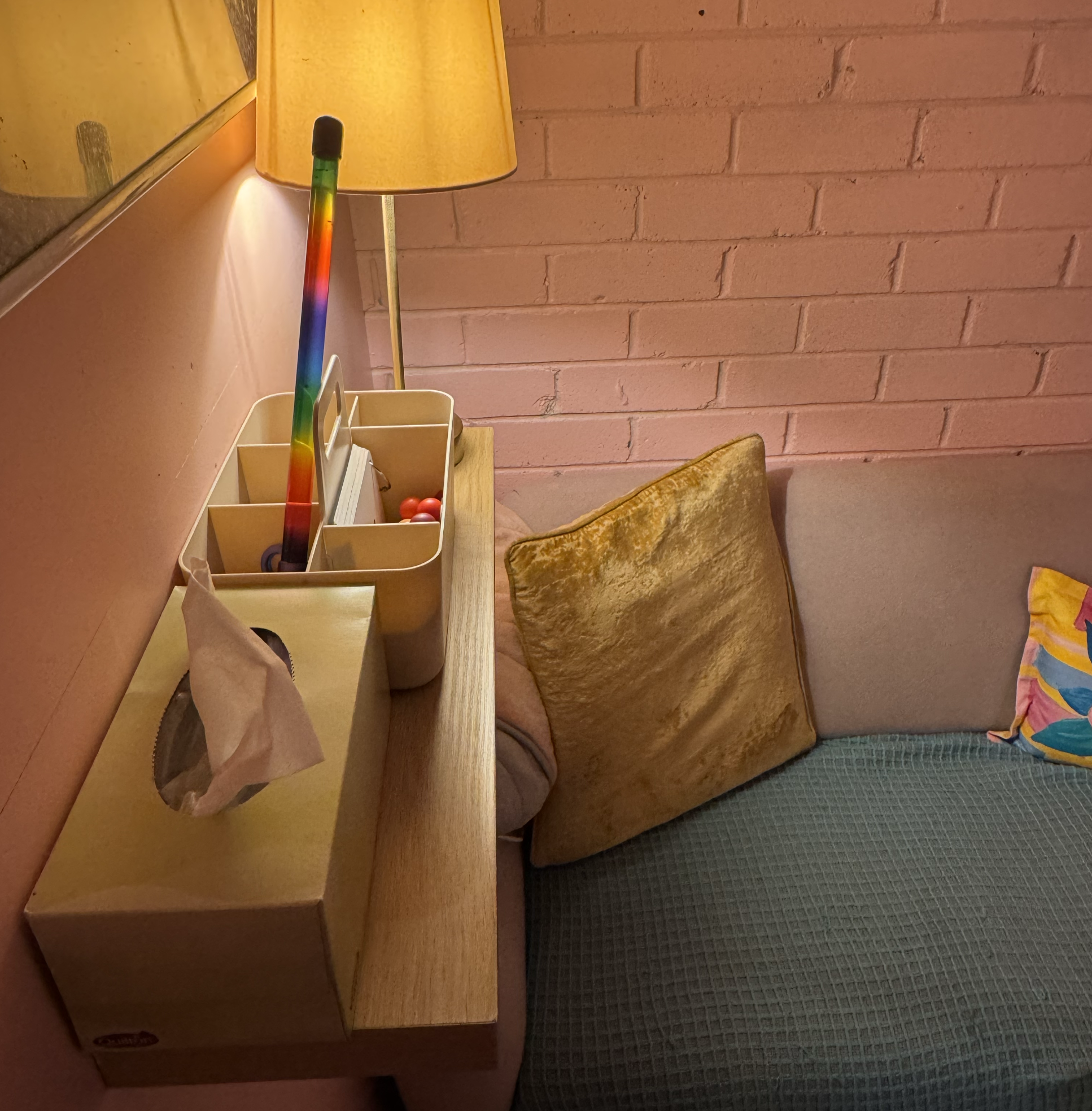 A corner of a room with a pink brick wall, a wooden desk with a beige organiser holding books and objects, and a chair with a worn, brown cushion. A nightstand with a golden lamp and a rainbow-colored crochet hook or decorative item leans against the wall. Partly visible colorful fabric on a bed or chair is in the lower right corner.