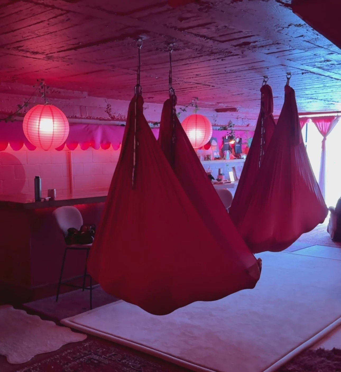 Decorated indoor space with hanging red fabric tents and pink lanterns, possibly set up for a celebration or event, with a small table and chairs in the background.