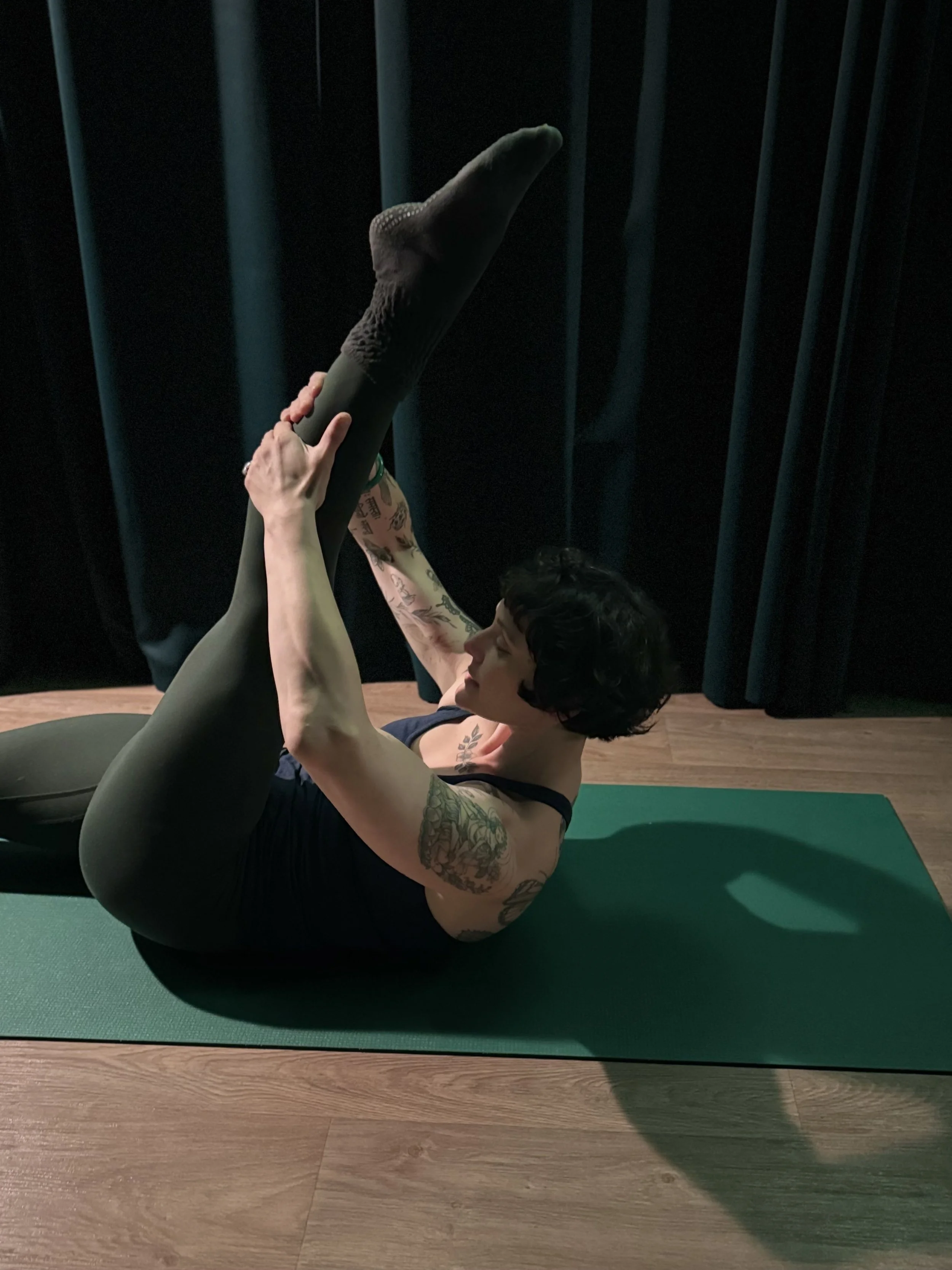 A woman with tattoos practicing yoga on a green mat, performing a pose with one leg raised and held by her hands in a dimly lit room with dark curtains.
