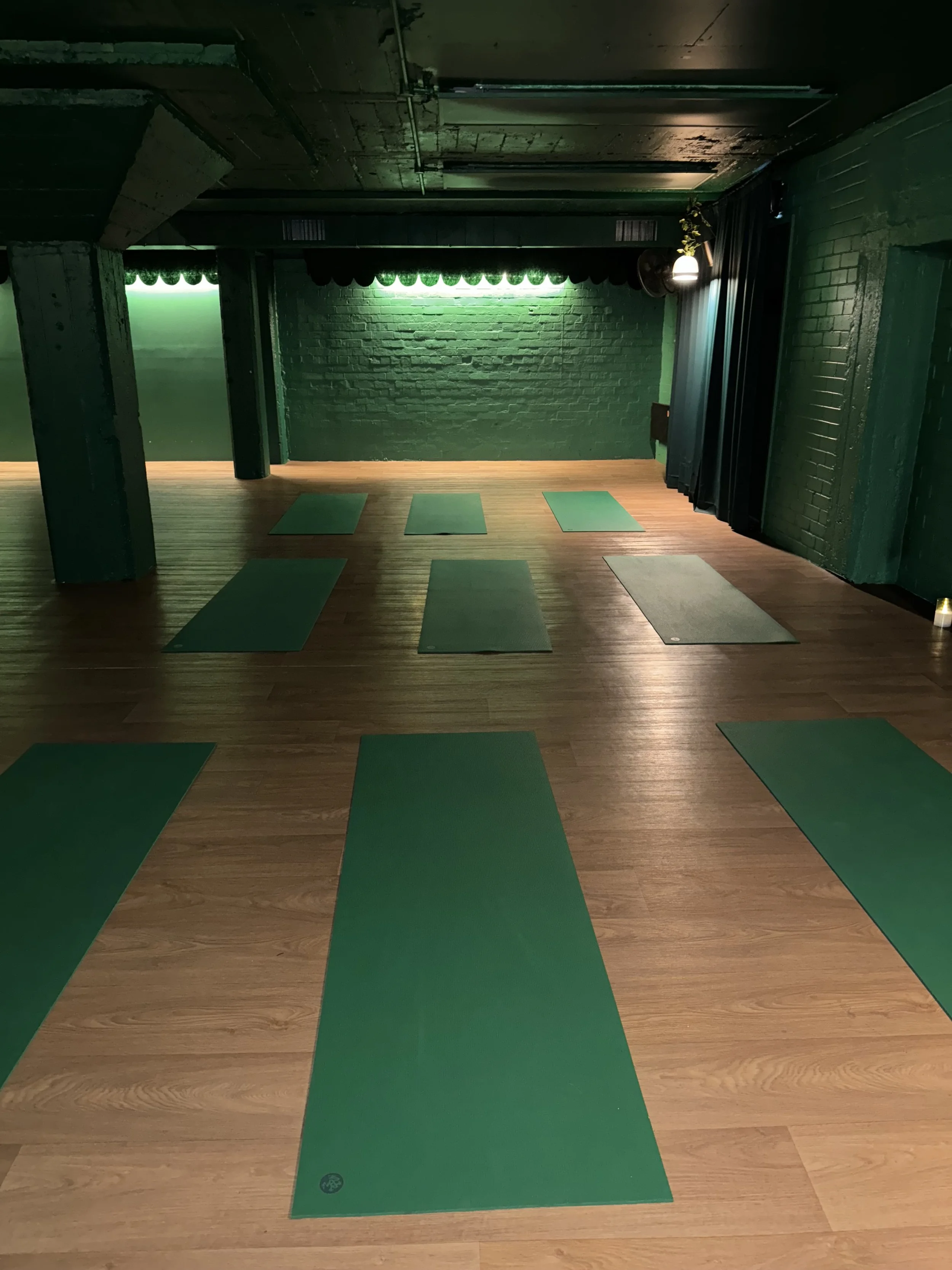An indoor yoga studio with wooden flooring, a black brick wall, and green yoga mats laid out on the floor.
