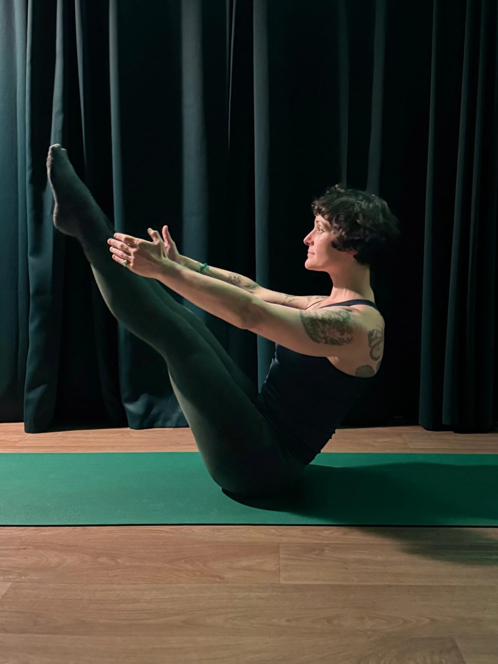 A woman with tattoos practicing yoga indoors on a green mat, sitting on her sit bones with legs extended upwards and reaching her feet with her hands, in front of black curtains.