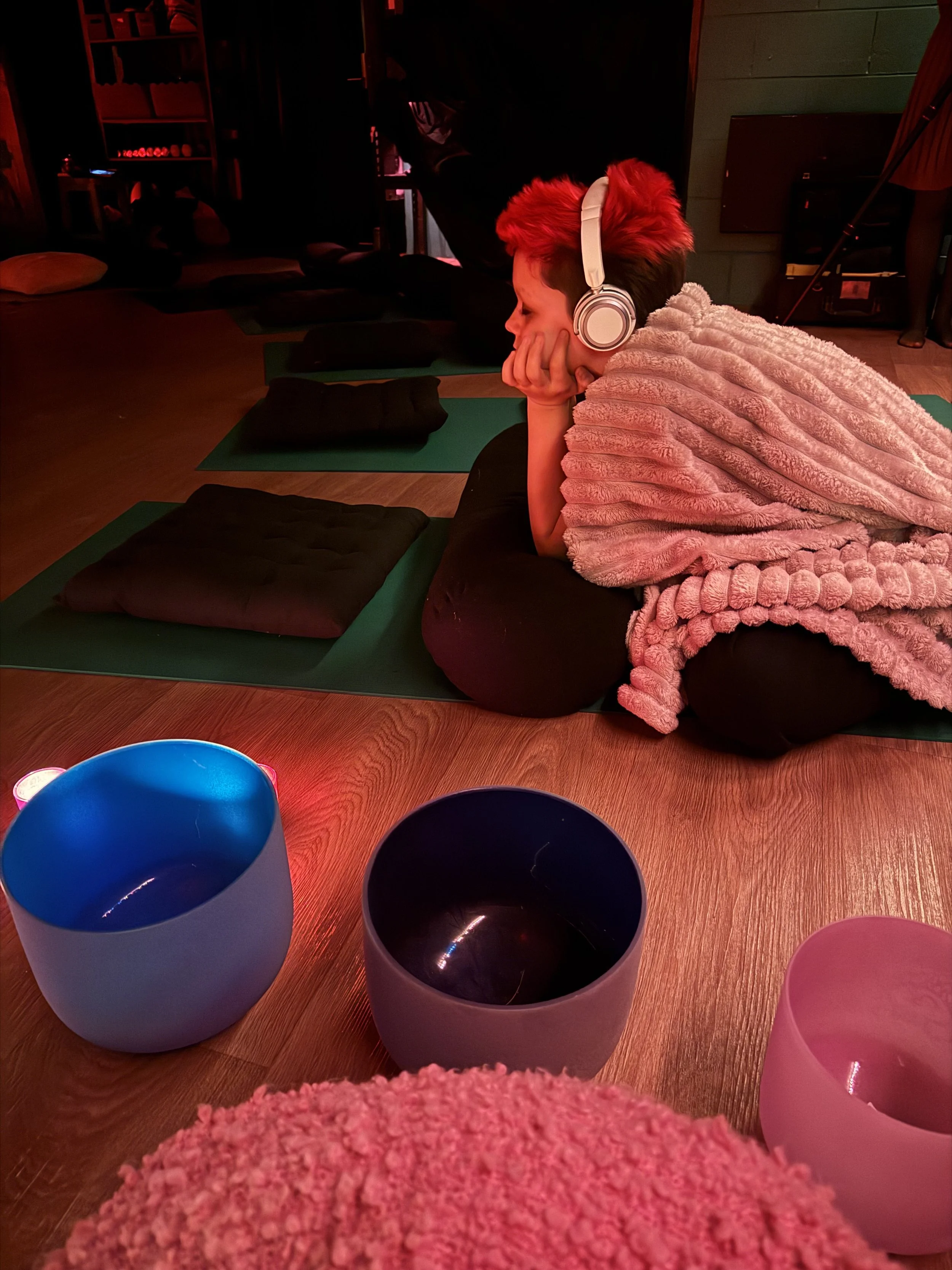 A young person with bright red hair wearing headphones and a fluffy pink blanket is lying on their stomach on a yoga mat, resting their chin on their hand, in a dimly lit room with cushions and colorful bowls in the foreground.
