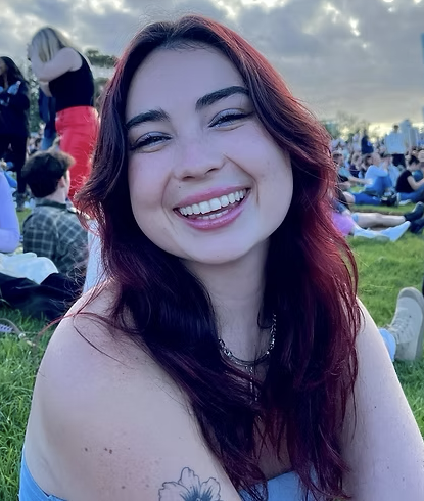 A woman with red hair smiling at an outdoor event with many people sitting on the grass and cloudy sky in the background.