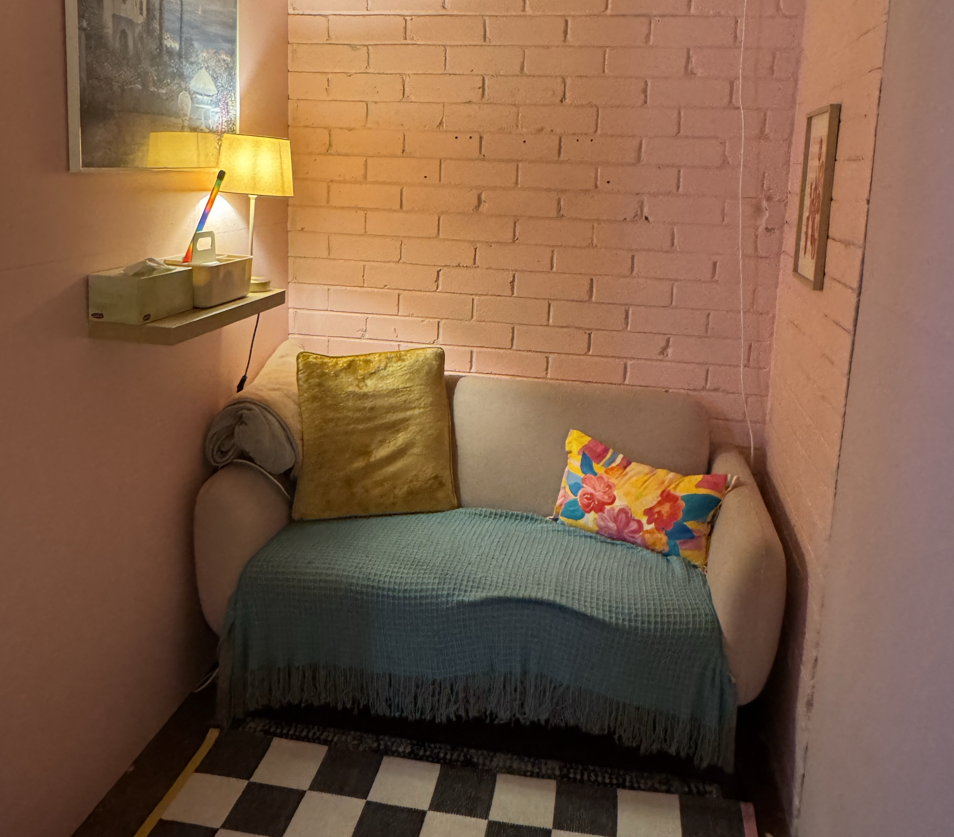 Corner of a room featuring a pink brick wall, part of a beige sofa with a velvety gold pillow, black and white checkered patterned floor, and a small wooden shelf with a lamp and tissue box. There is also a framed picture on the wall.