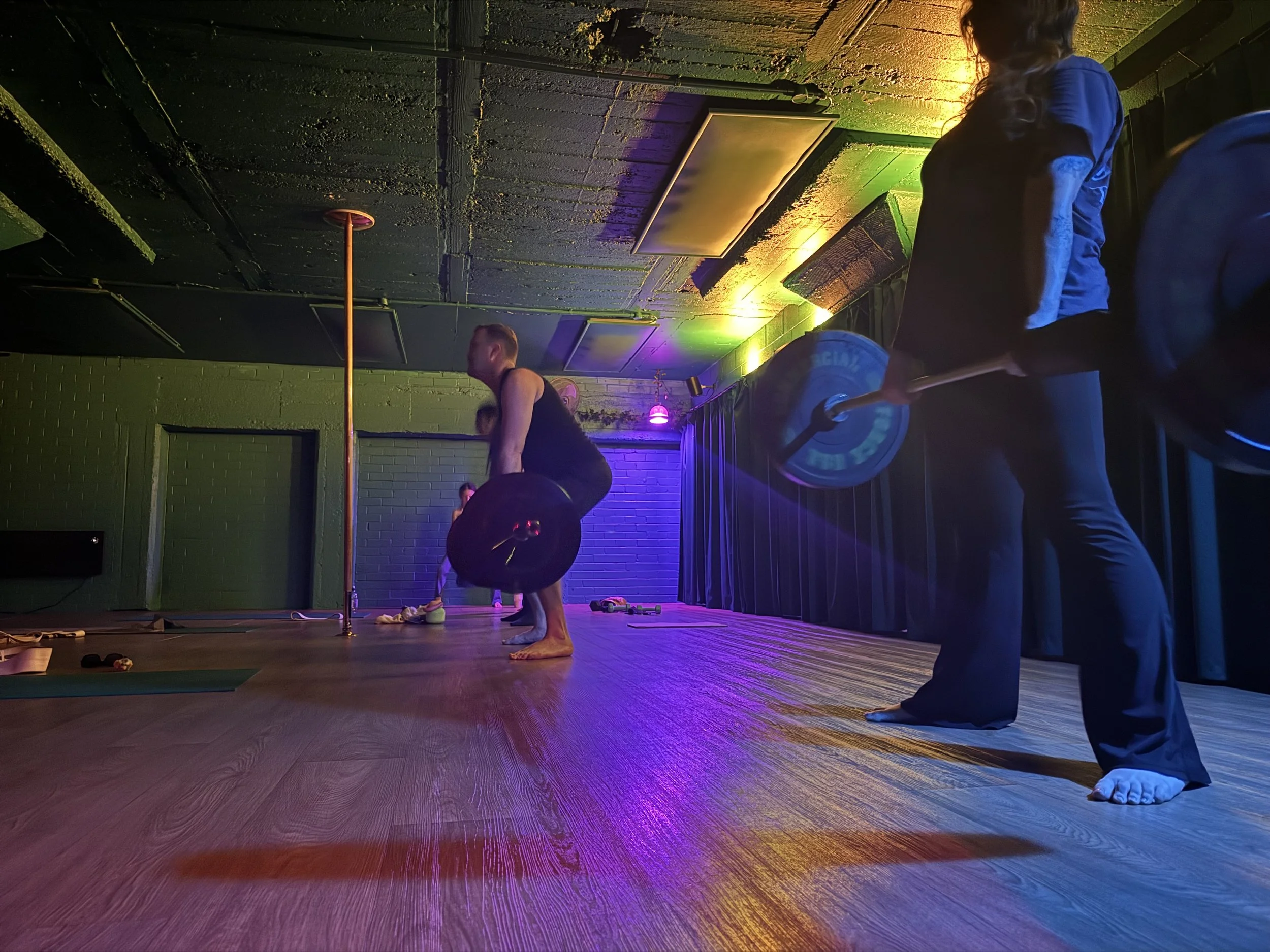 People practicing yoga in a dimly lit studio with colorful LED lights, some in a kneeling or bending pose, on a wooden floor.