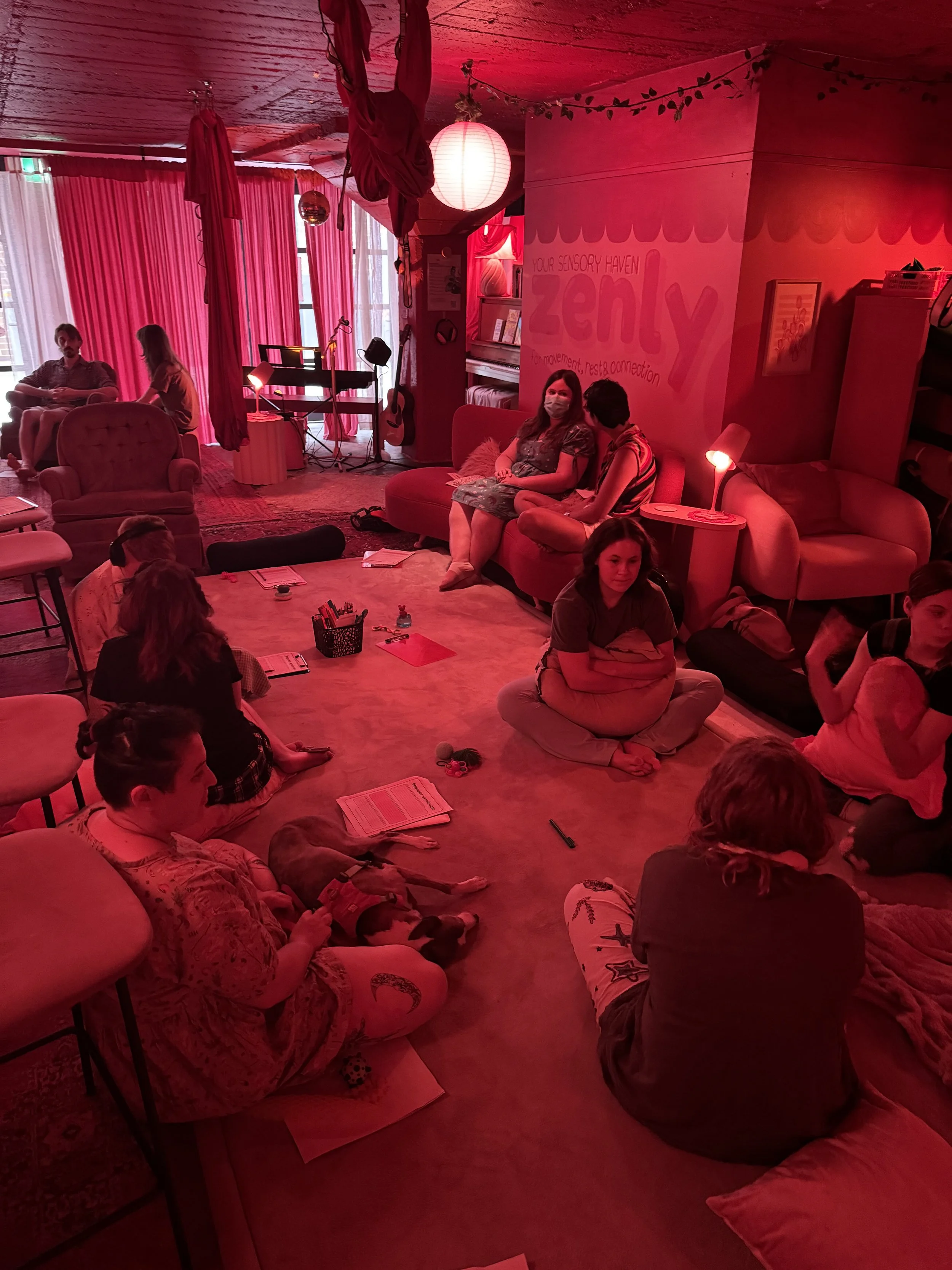 People sitting in a cozy, dimly lit room with red lighting, some sitting on couches and floor, some engaging in conversation, with musical instruments and books visible in the background.
