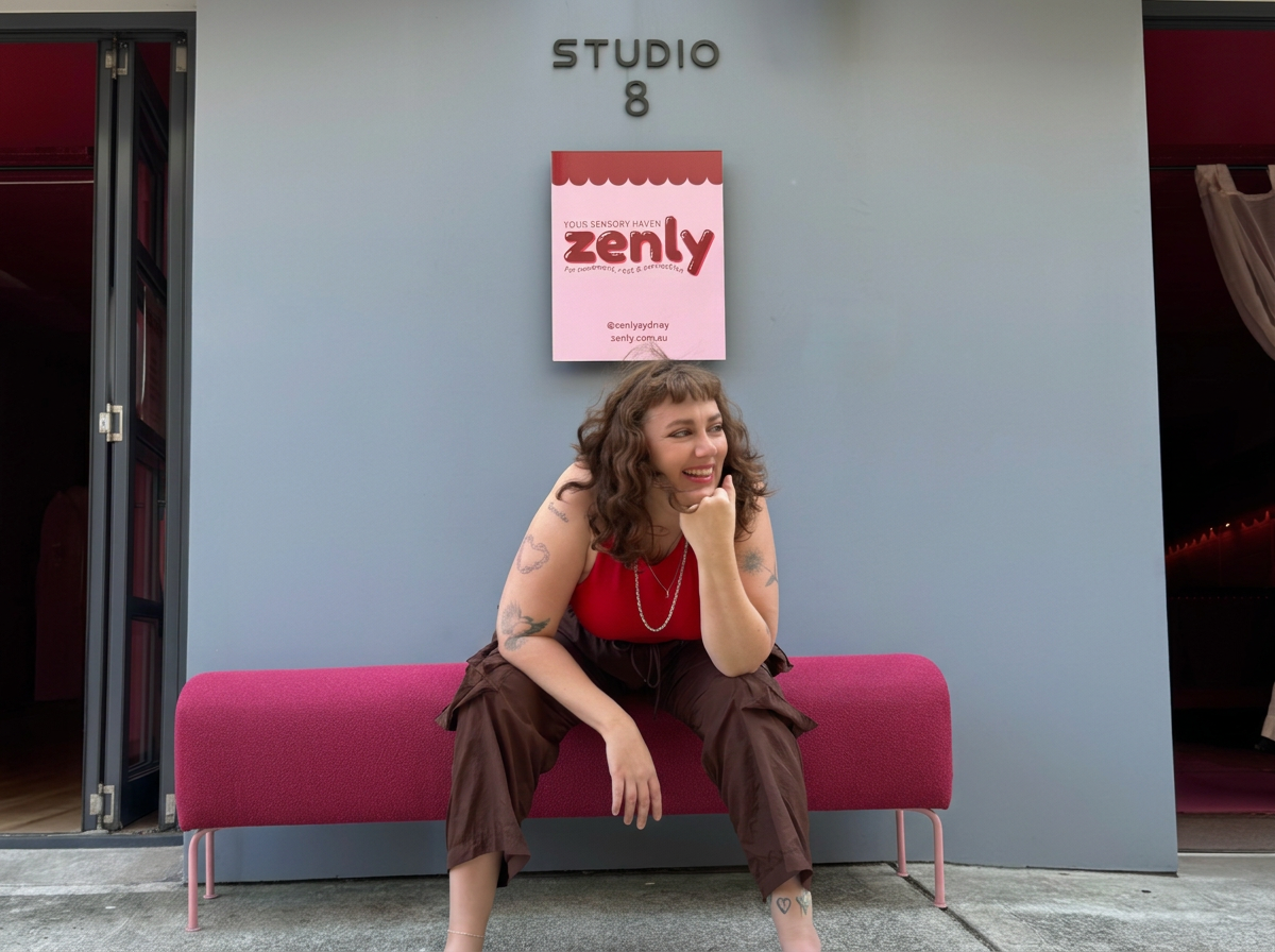 A woman sitting on a pink bench outside a building, smiling and resting her chin on her hand, with a sign that reads 'Zenly' and a door labeled 'Studio 8' behind her.