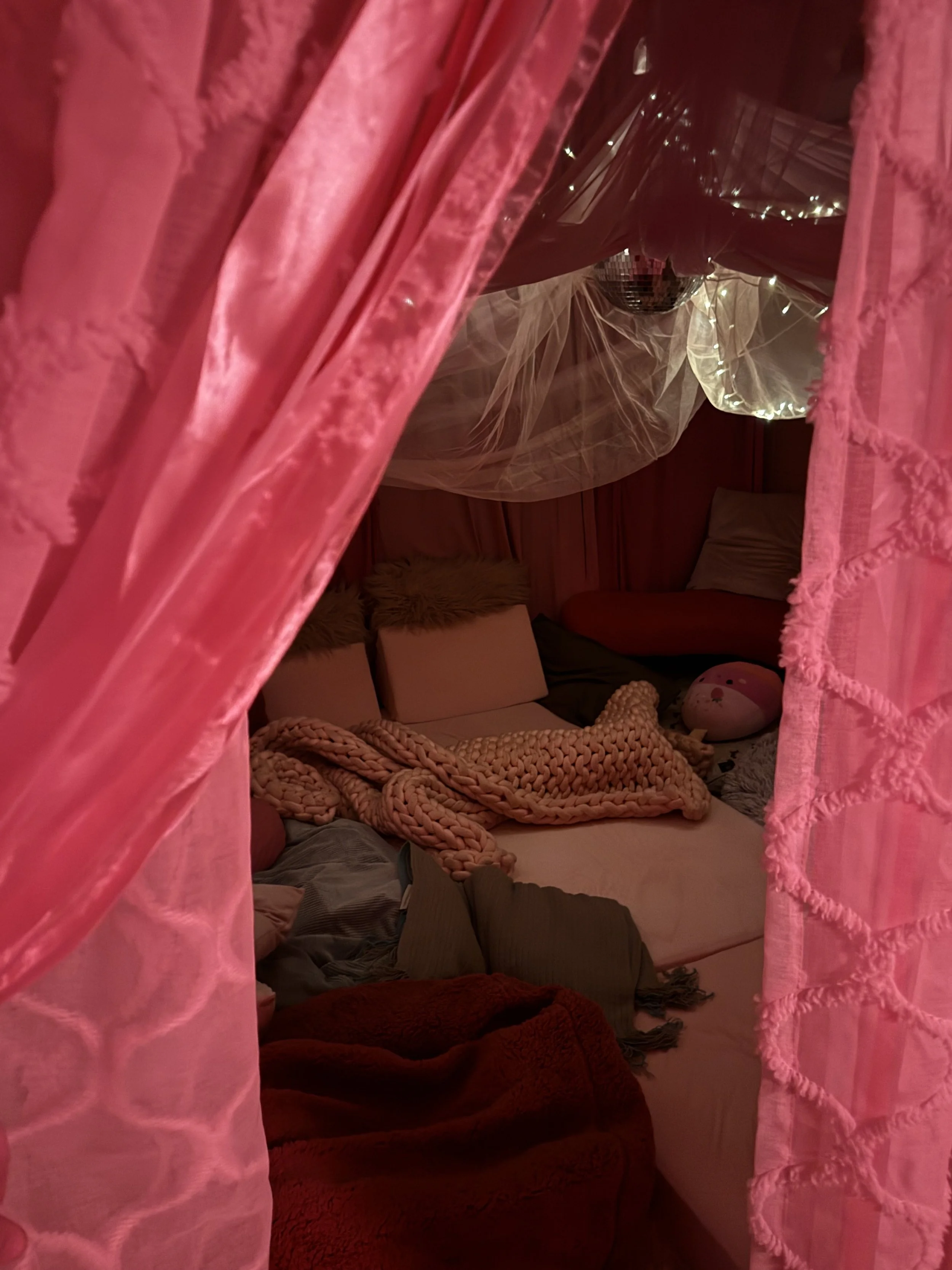 Looking into a cozy room through pink curtains, with a bed covered in blankets, pillows, and a red plush blanket. There are string lights and a disco ball in the background.