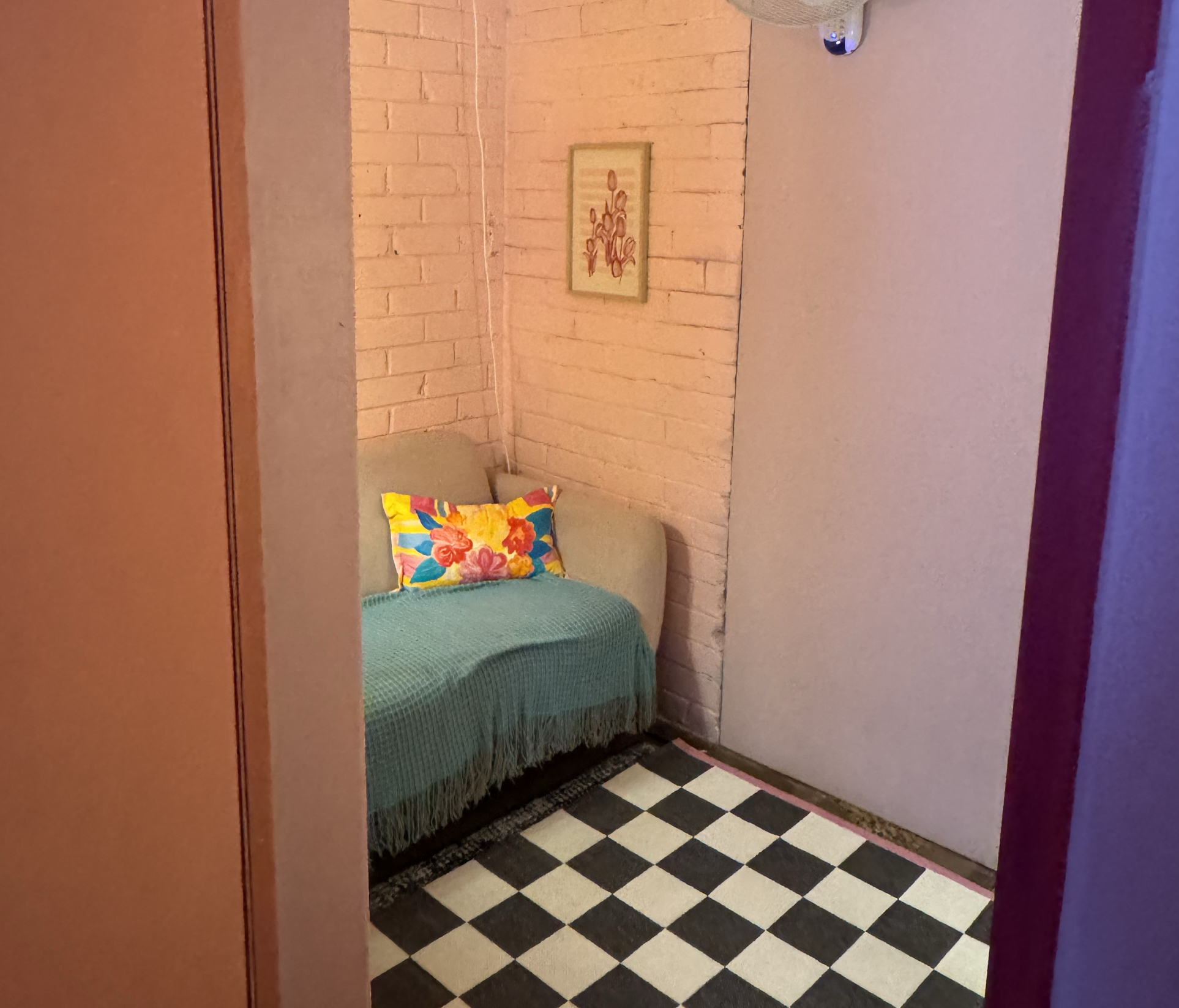 A cozy bedroom with a bed covered in a green quilt and a colorful floral pillow, a beige headboard, a framed artwork of flowers on the exposed brick wall, and checkered black and white flooring.