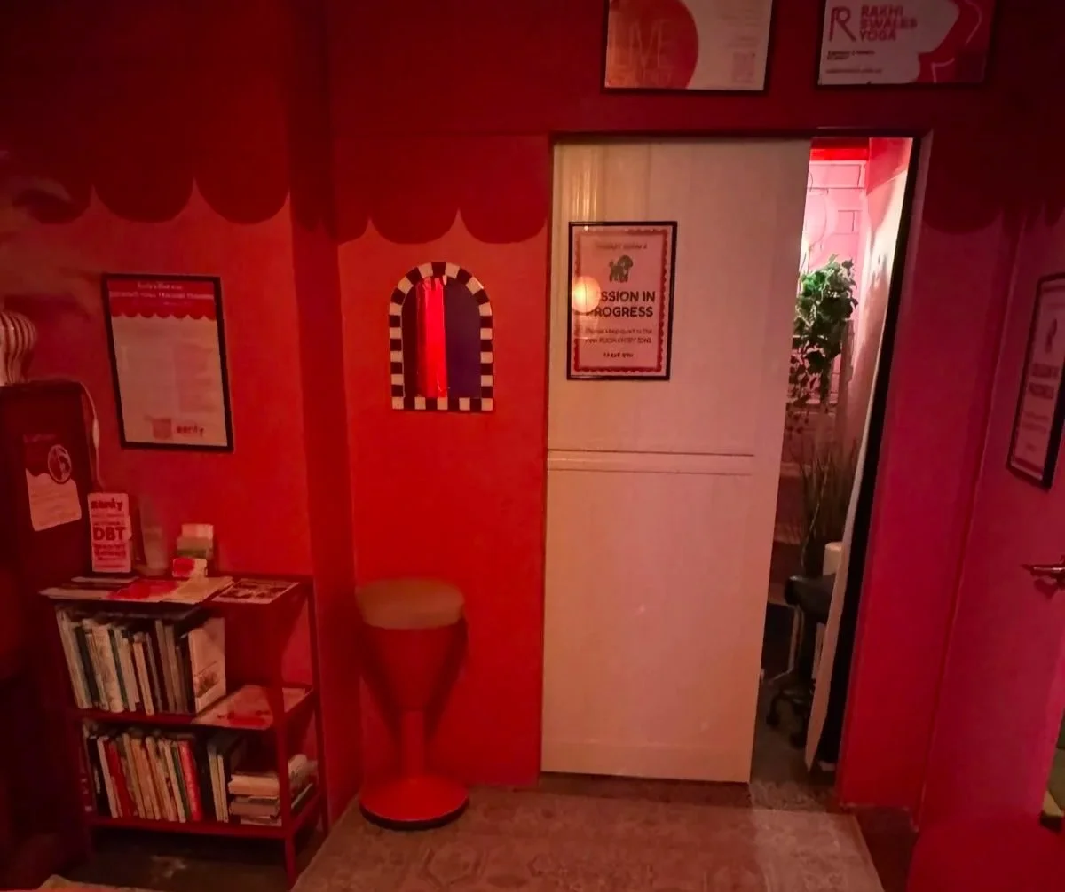 Red-walled interior with framed artwork, a bookshelf filled with books, a small table with items on top, a doorway leading into a room with a plant, and vintage-style red and black striped mirror.