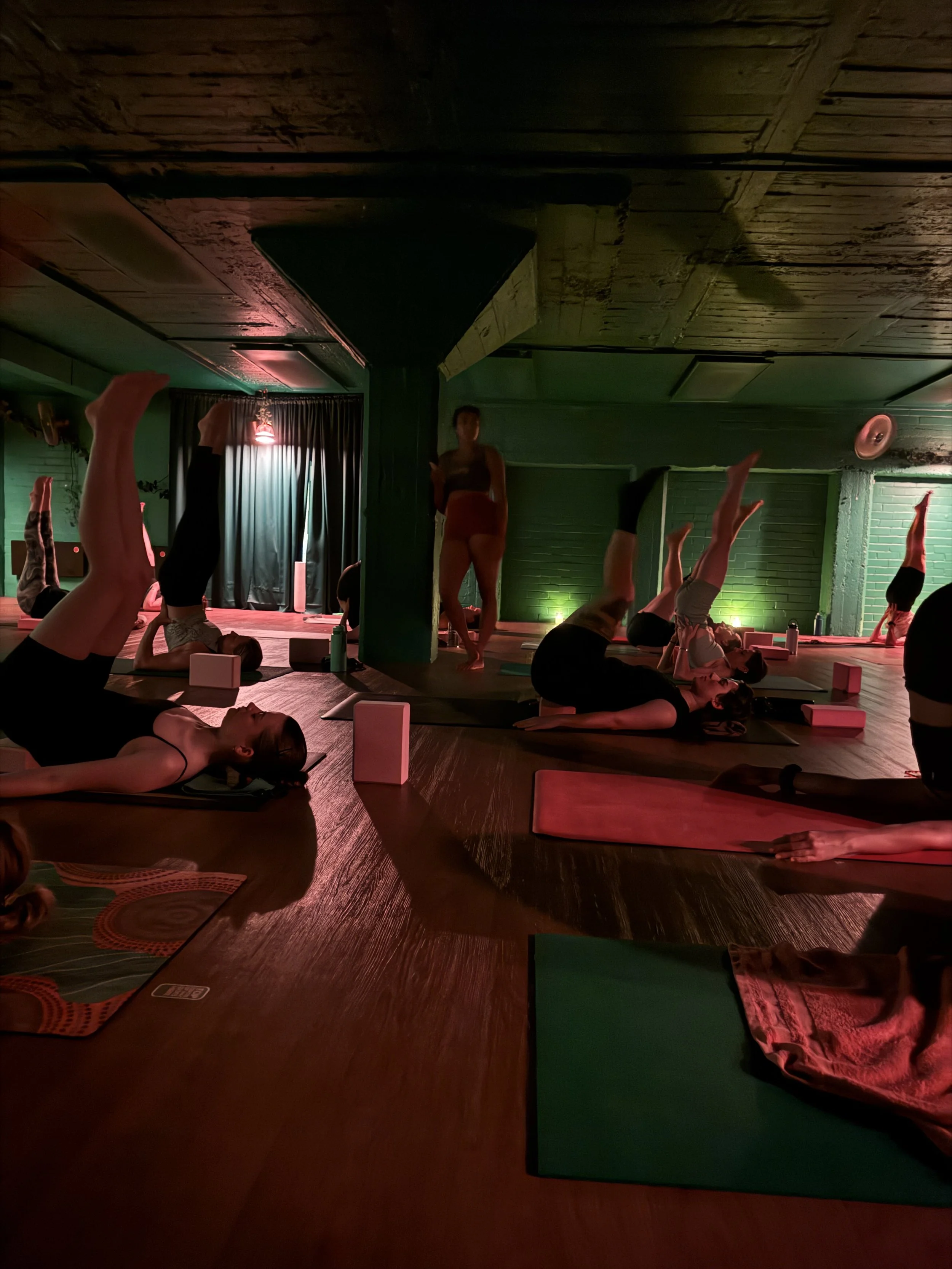 People practicing yoga in a dimly lit room with green walls, black curtains, and a wooden ceiling, using yoga mats and blocks.