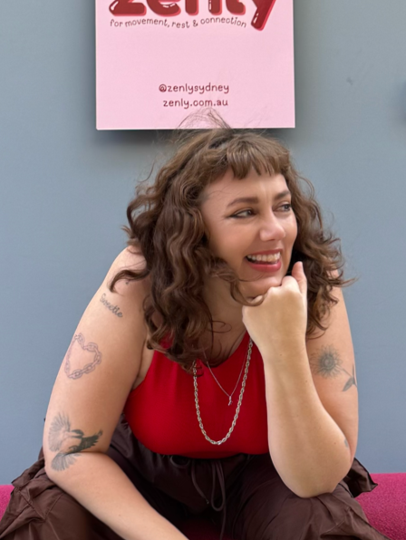Young woman with curly brown hair and tattoos on her arm, wearing a red top and a chain necklace, sitting and smiling with her hand on her chin, in front of a light blue wall with a pink sign above her.