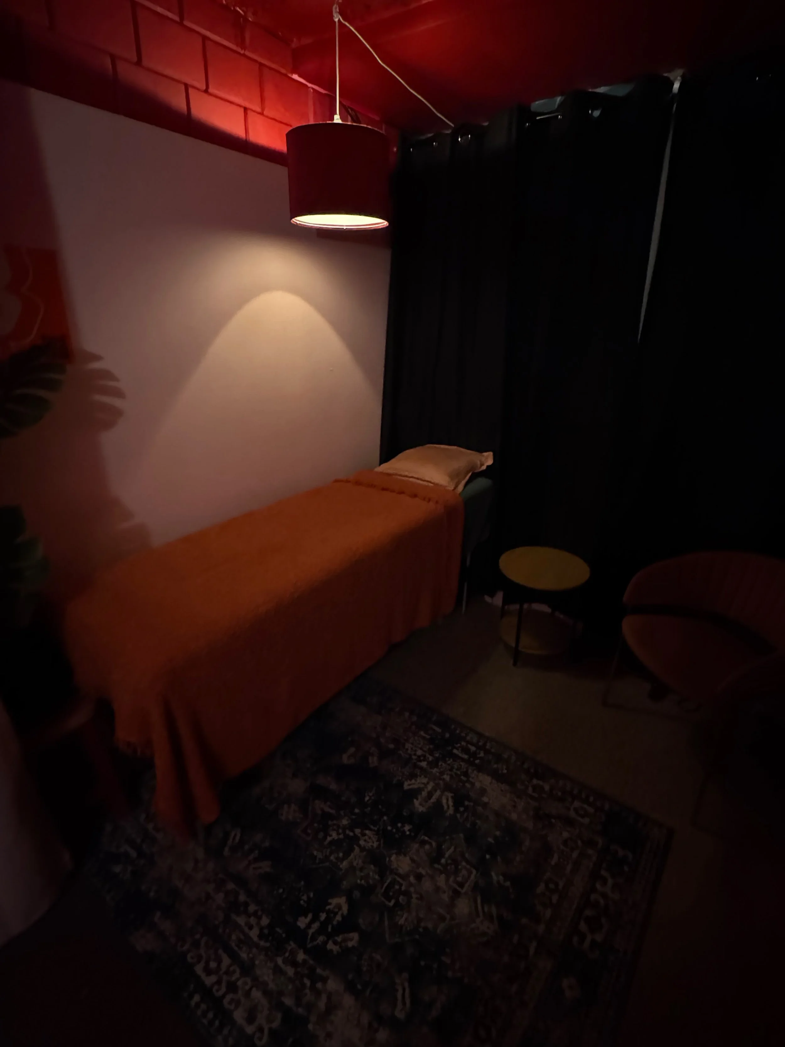 A dimly lit bedroom with a bed covered in an orange bedspread, a pillow, a patterned area rug, a small yellow side table, a black curtain, and a wall-mounted red lampshade casting a warm glow.