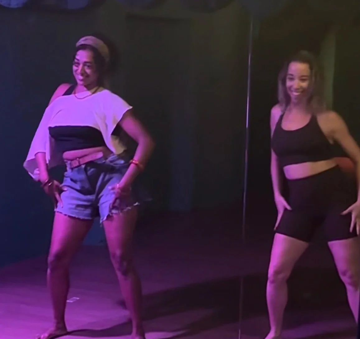 Two women dancing in a dimly lit room with pink and purple lighting, one wearing a white and black crop top with denim shorts, and the other wearing a black crop top and shorts.