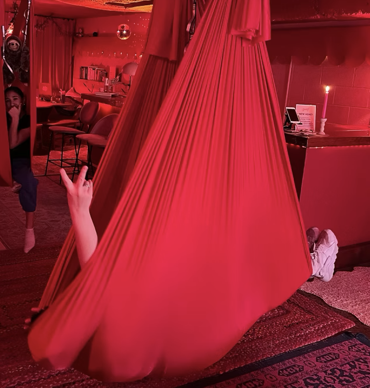 A person lying inside a red fabric hammock in a cozy, dimly lit room with red lighting. They are making a rock and roll hand gesture with their hand and are wearing white sneakers. The room has a pink candle, a small table with a phone, and a chair. There is a woman with long hair and a black shirt standing nearby, smiling. The background includes chairs, books, and a bookshelf, creating a warm, inviting atmosphere.