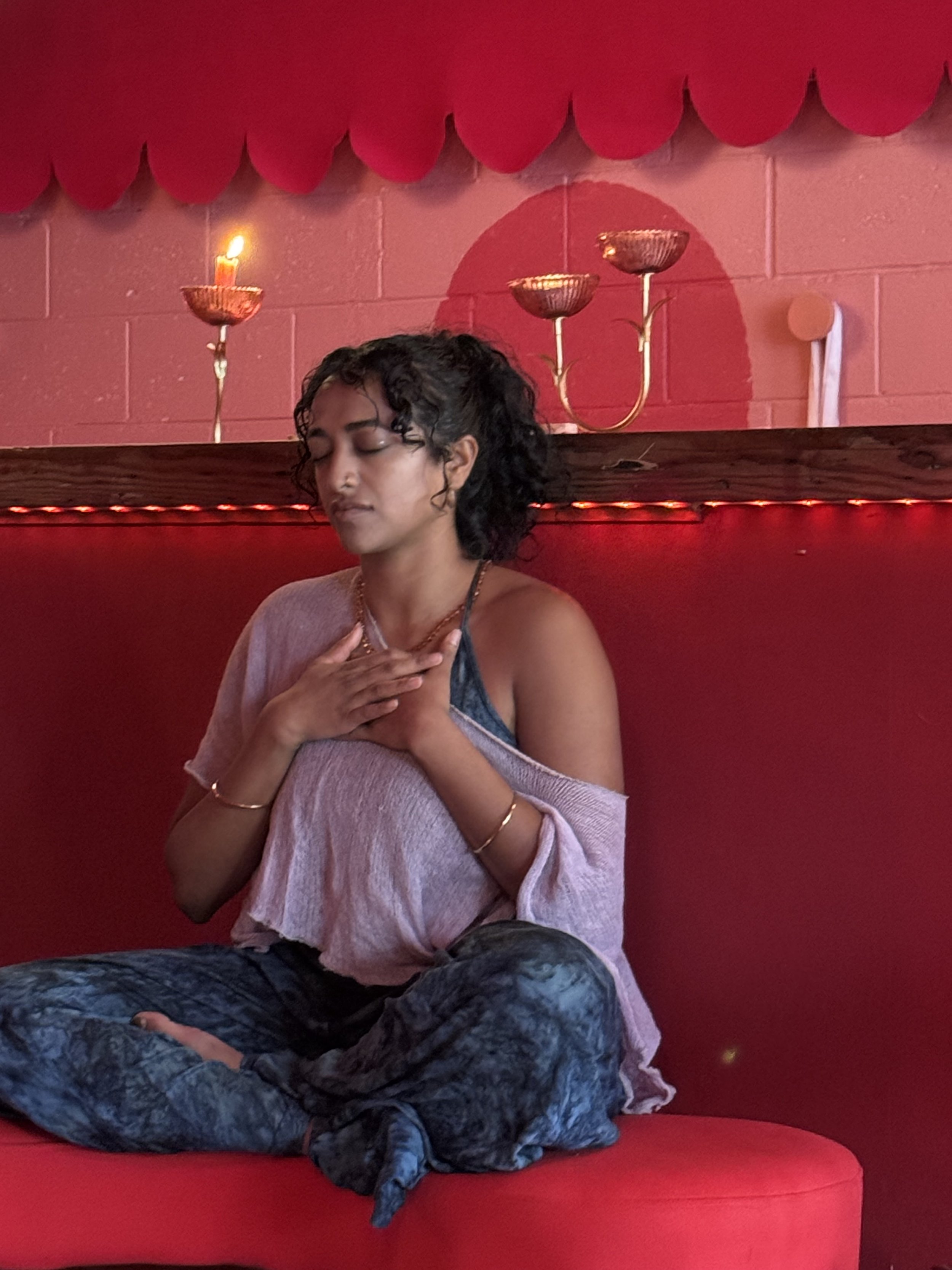 A woman with curly hair, closed eyes, and a peaceful expression, sitting on a red cushioned seat in a room with red walls, a red scalloped decorative border, golden wall-mounted candle holders, and a lit candle.