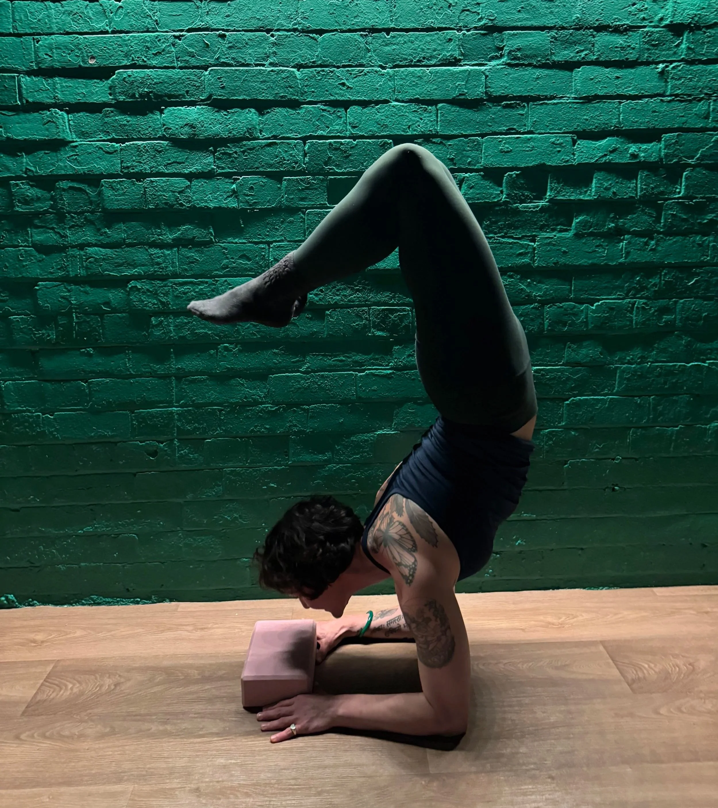 A person practicing yoga in a forearm stand against a green brick wall, with a yoga block supporting their forearms.